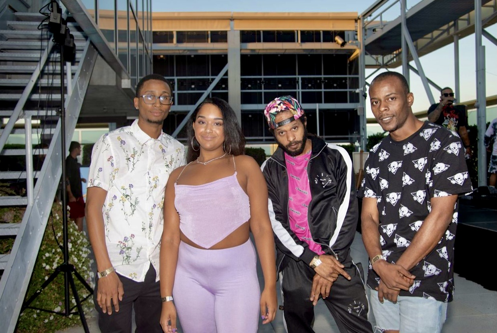 Four people standing outdoors in front of a building with glass and metal structures, posing for a photo.