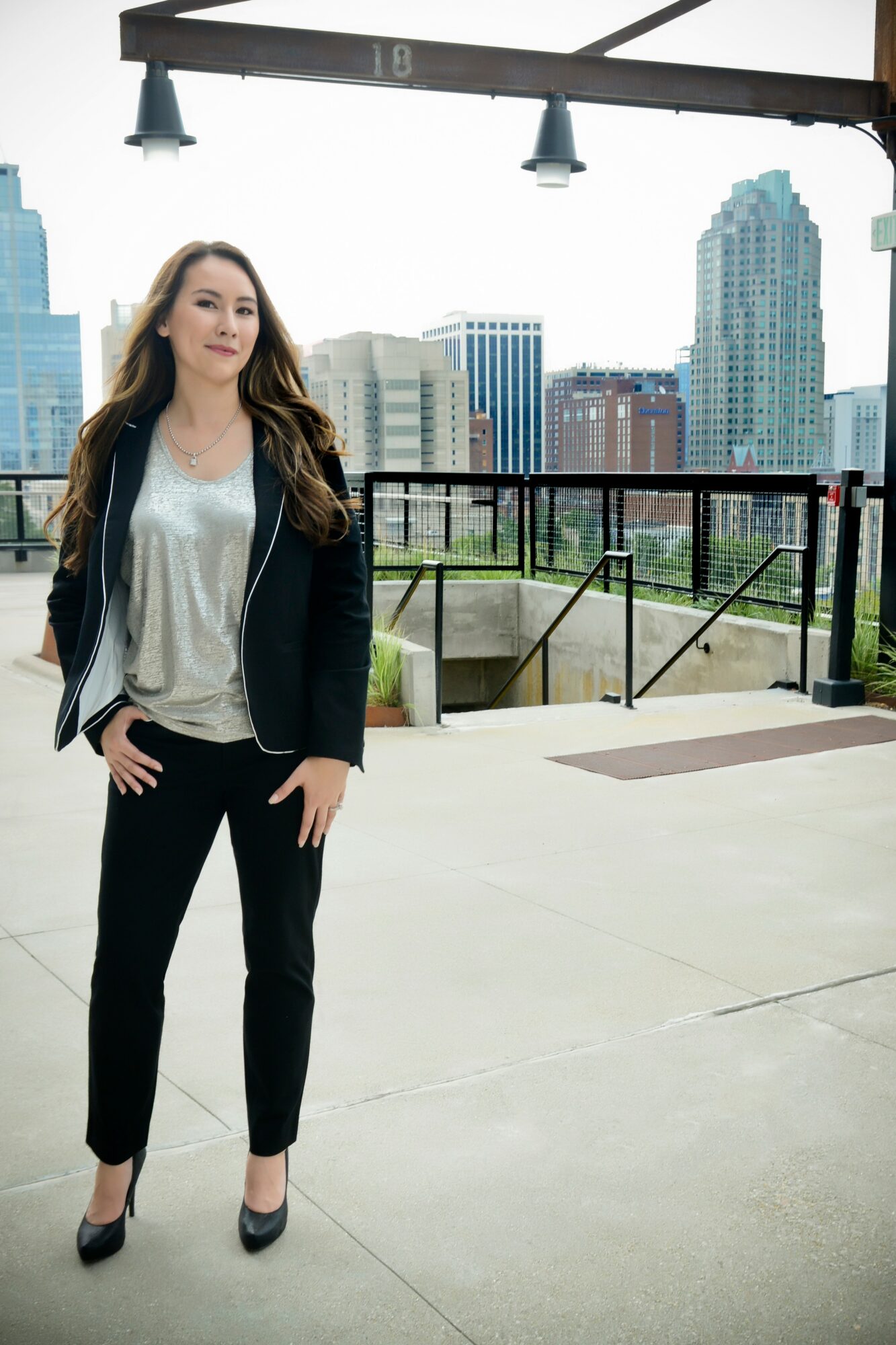Woman standing outdoors on a balcony with city skyline in background, wearing black pants and blazer, smiling.