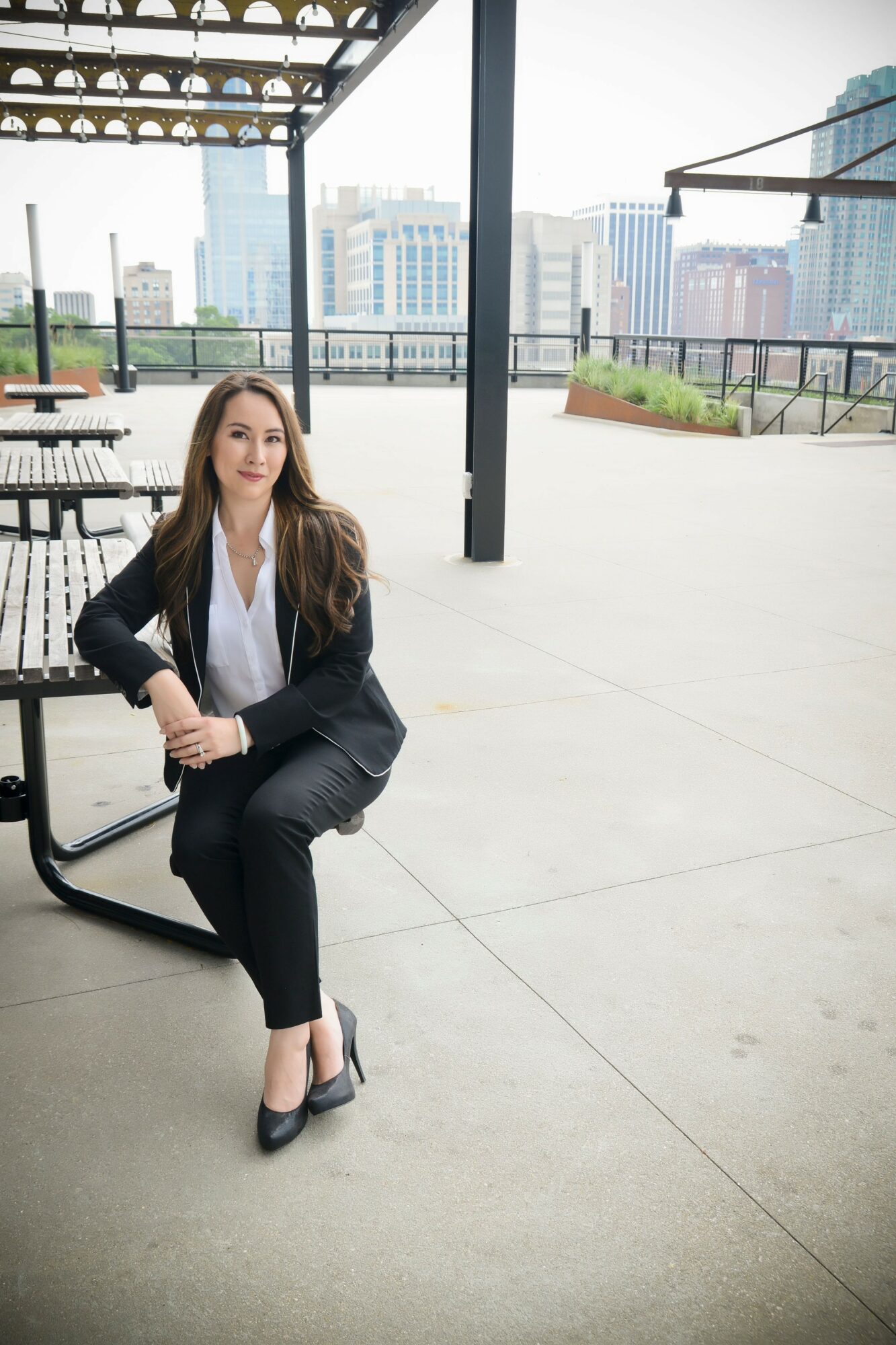 Woman sitting on a chair outdoors with city buildings in the background, under a shaded structure.