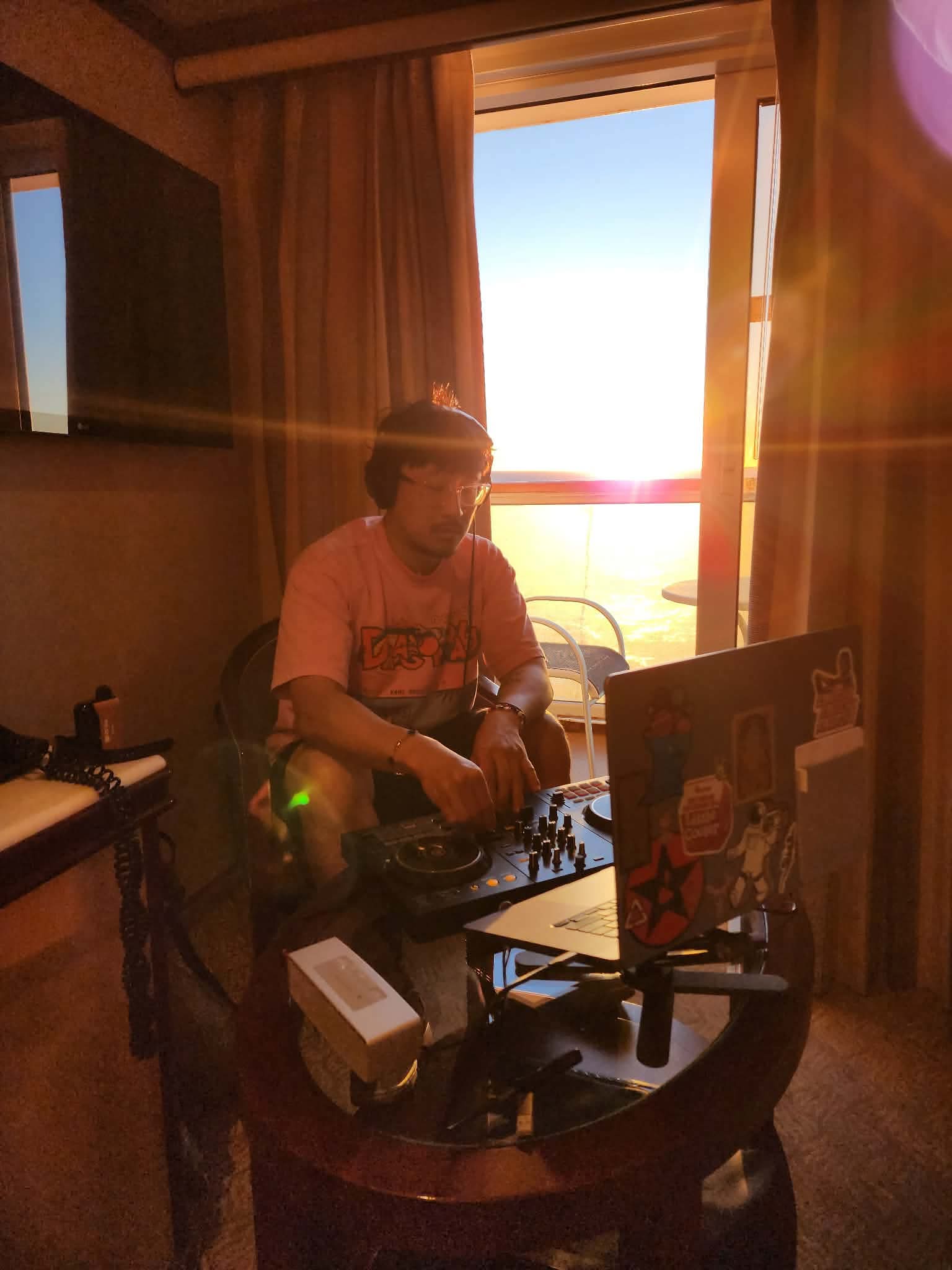 Person wearing glasses and a pink shirt stands at a DJ setup near a window with sunlight streaming in.