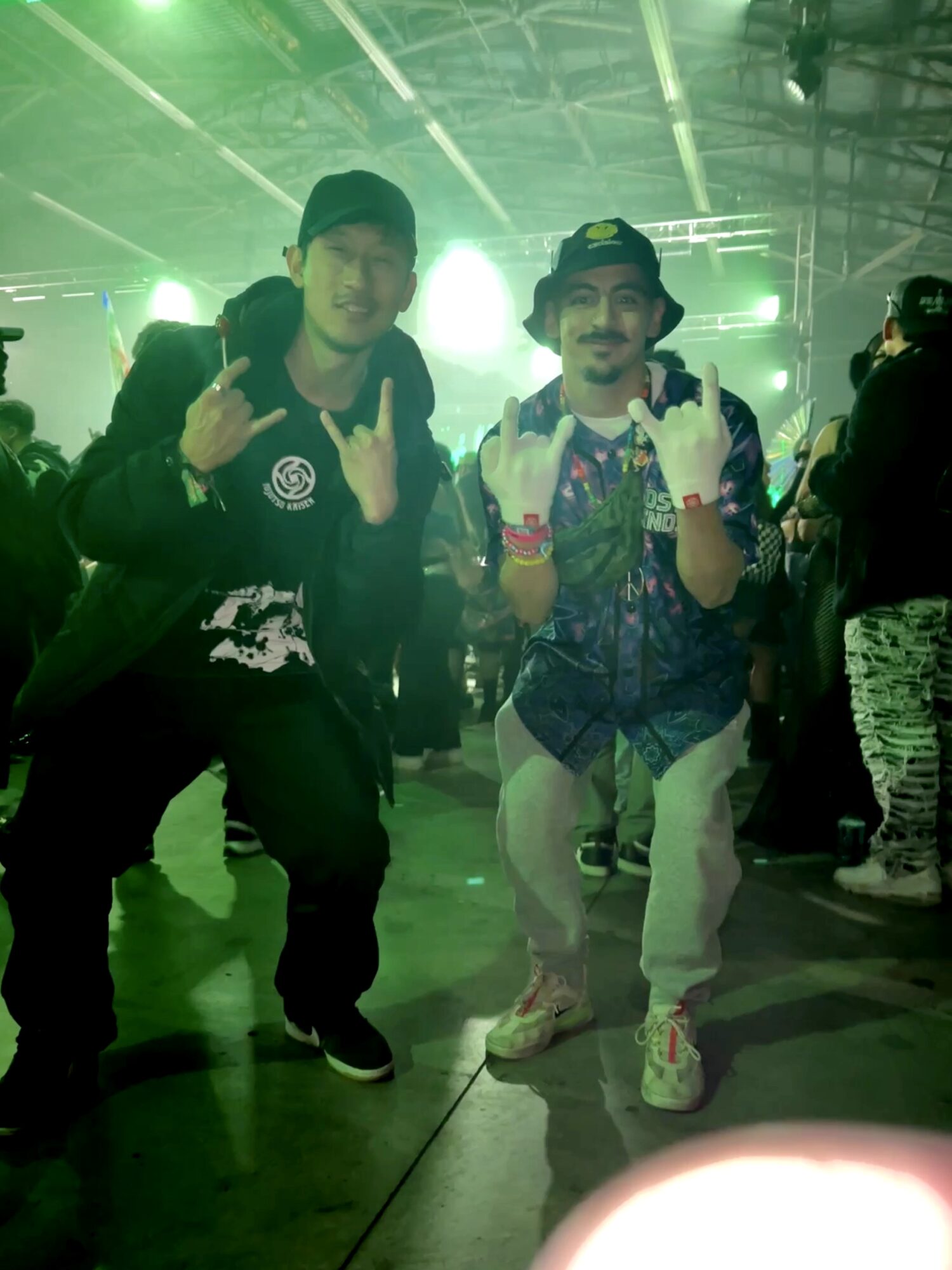 Two people posing at a concert or event, making hand gestures, with bright green stage lights in the background.