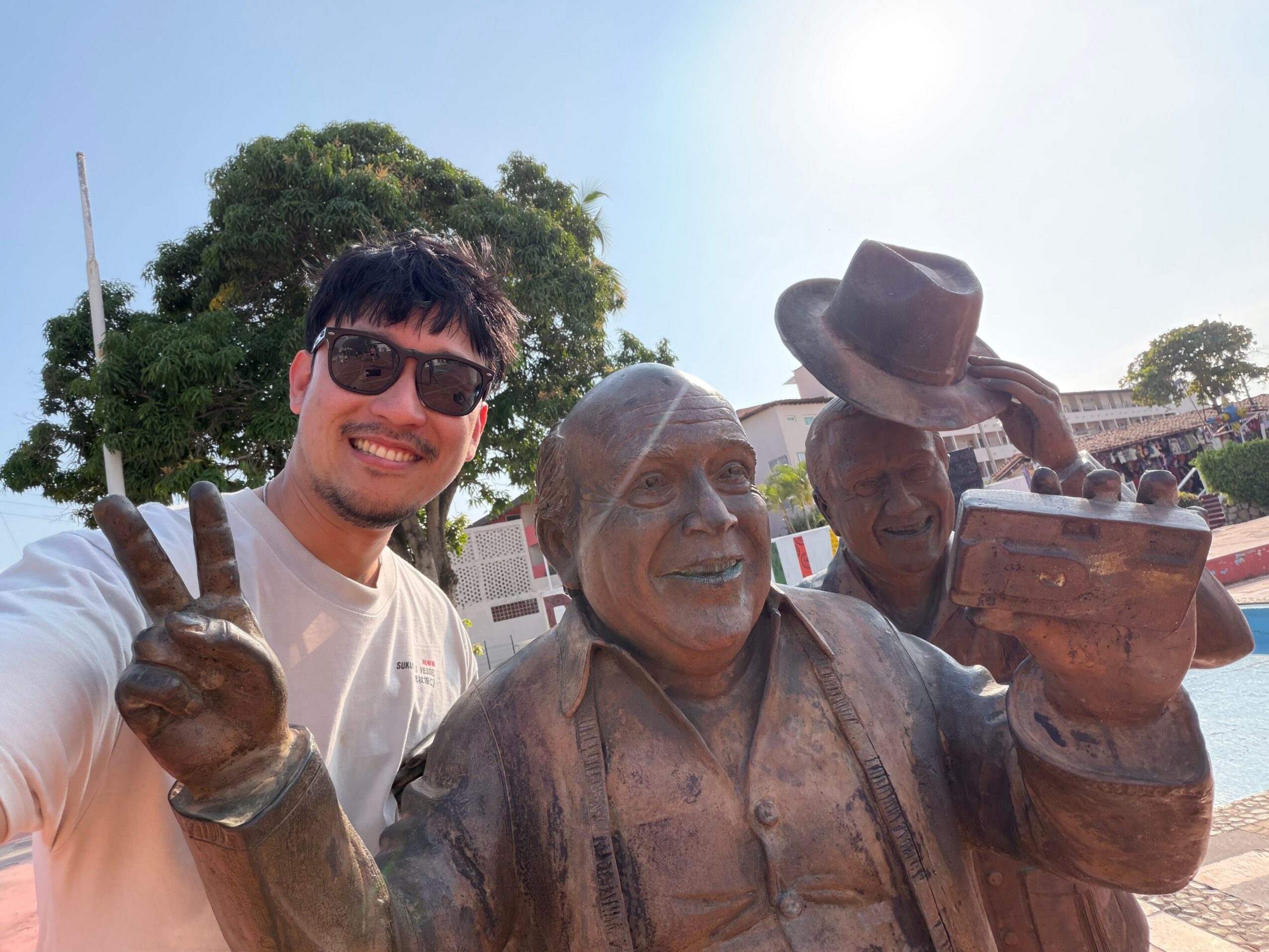 Young man with sunglasses smiling and making a peace sign next to a bronze statue of a man with a hat holding a book outdoors.