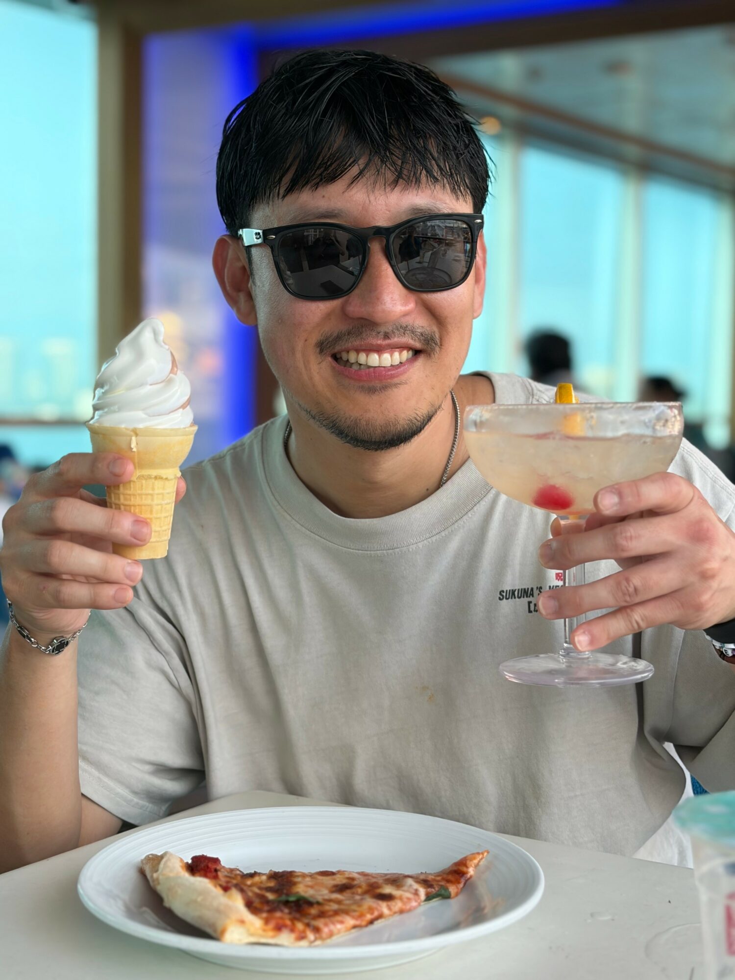 Young man wearing sunglasses smiling, holding ice cream cone and cocktail, with pizza on plate in front, indoors with large windows.