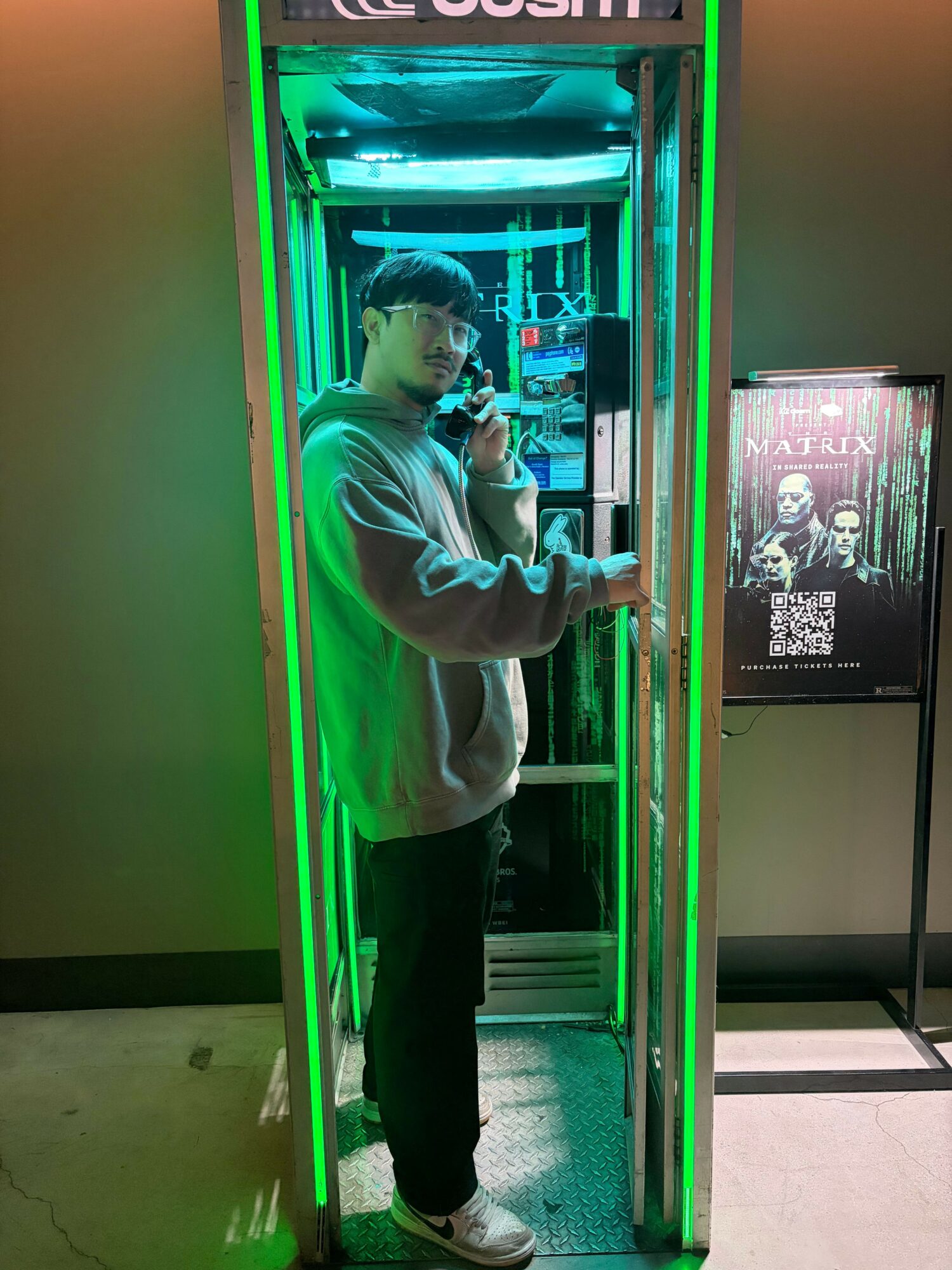 Person taking a selfie inside a green-lit phone booth, holding a phone and wearing a hoodie, with a QR code on the wall behind.