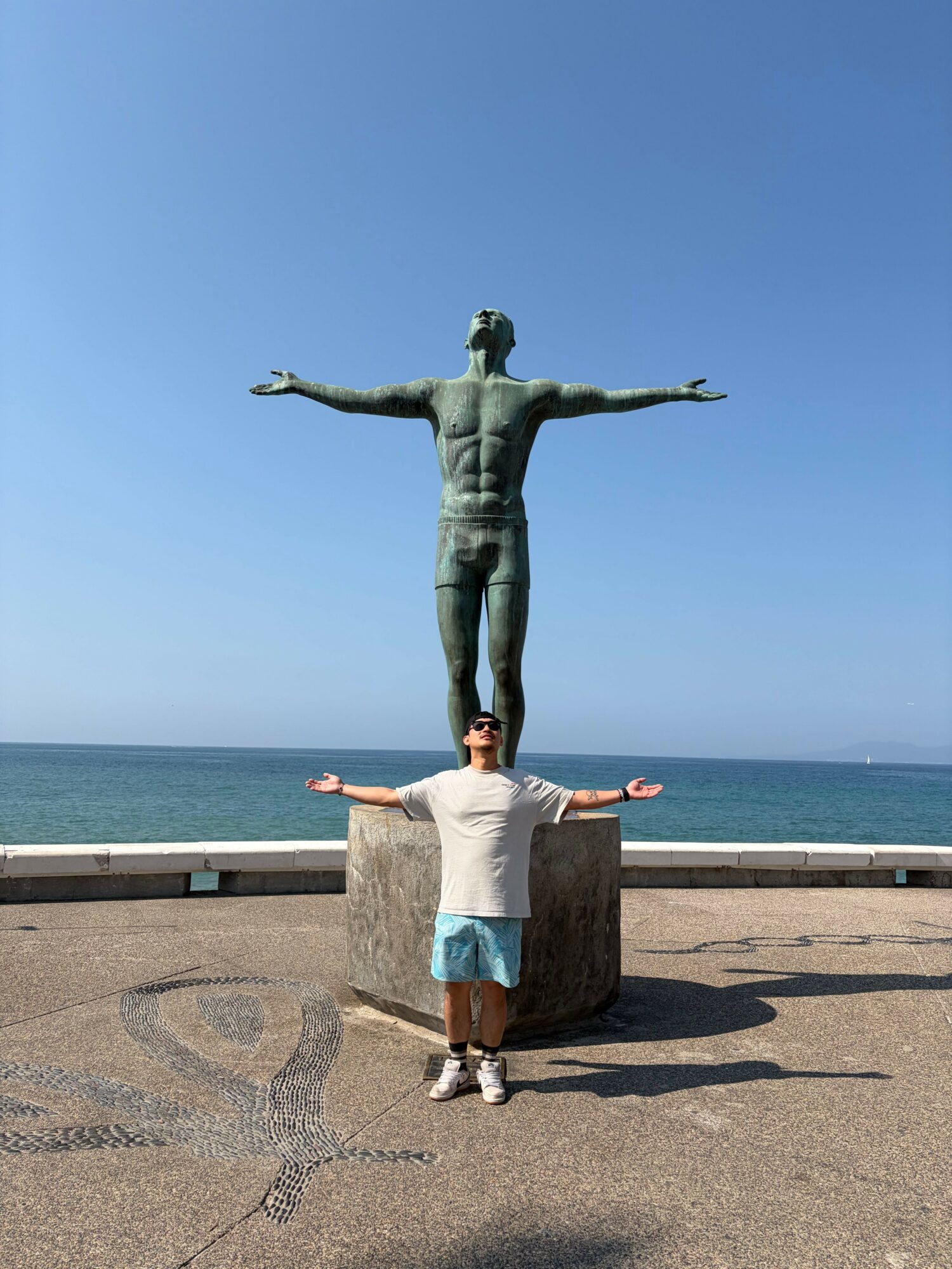 Person standing on a beach with arms outstretched, statue of Christ with arms open in the background, ocean and sky visible.