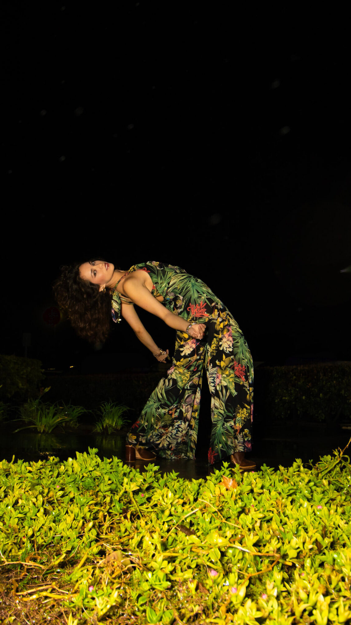 Person bending backward outdoors at night, wearing a floral outfit, with green plants in foreground.