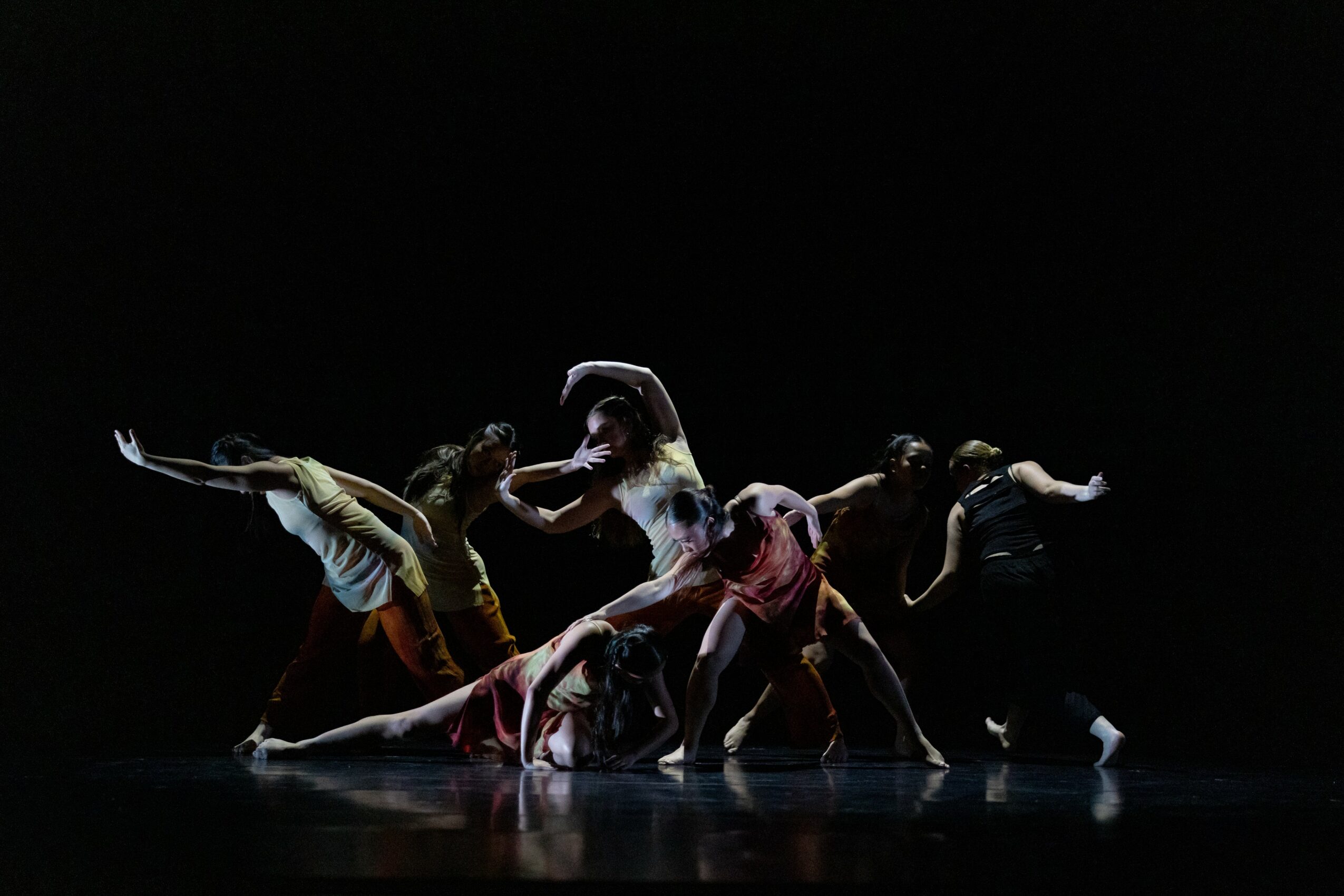 Group of dancers performing on stage with dark background, in various dynamic poses, illuminated by stage lighting.