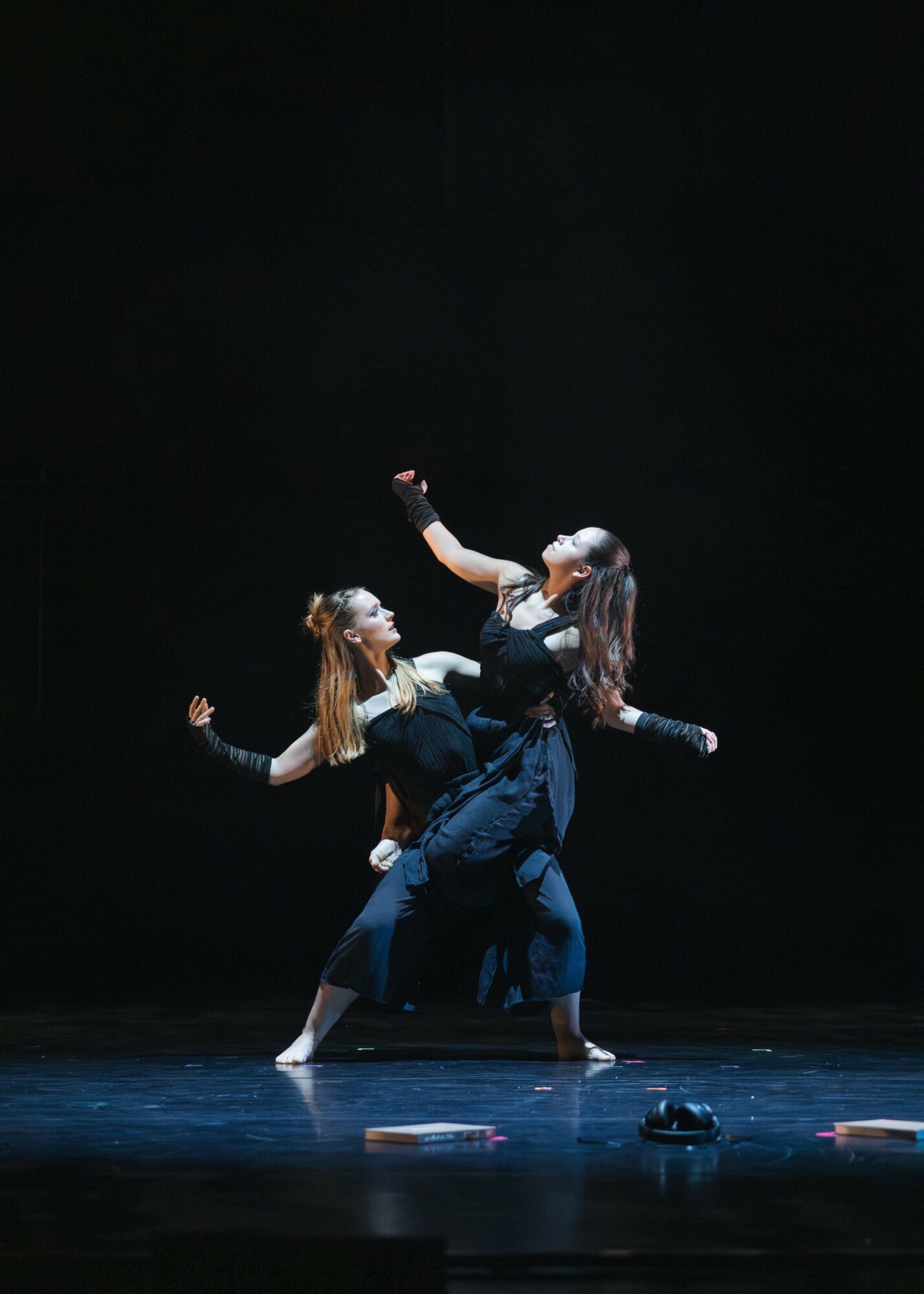 Three dancers perform on stage with a dark background, one lifted by the others, in a contemporary dance pose.
