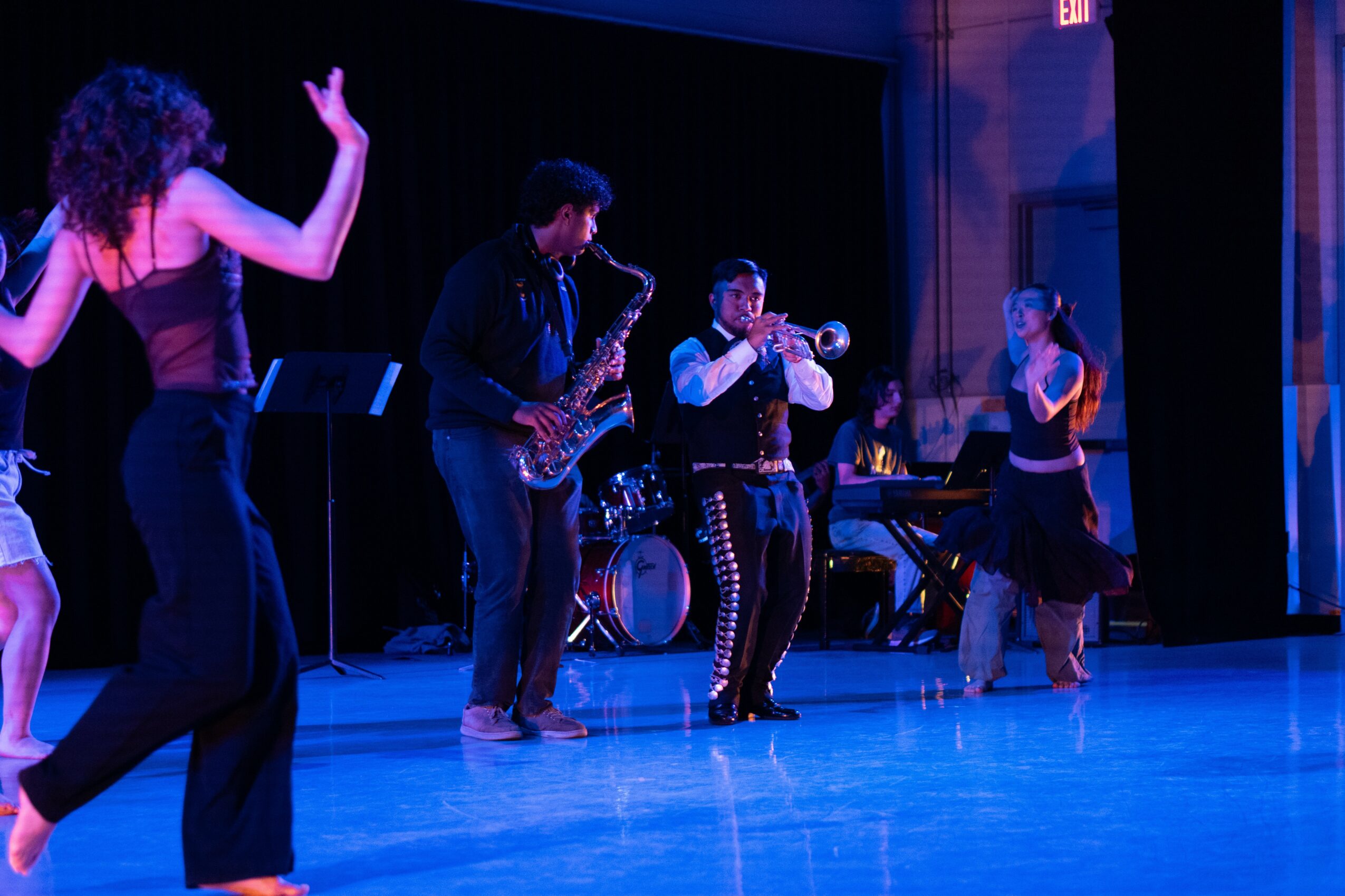 Group of five people dancing and playing instruments on stage with a dark background and blue lighting.