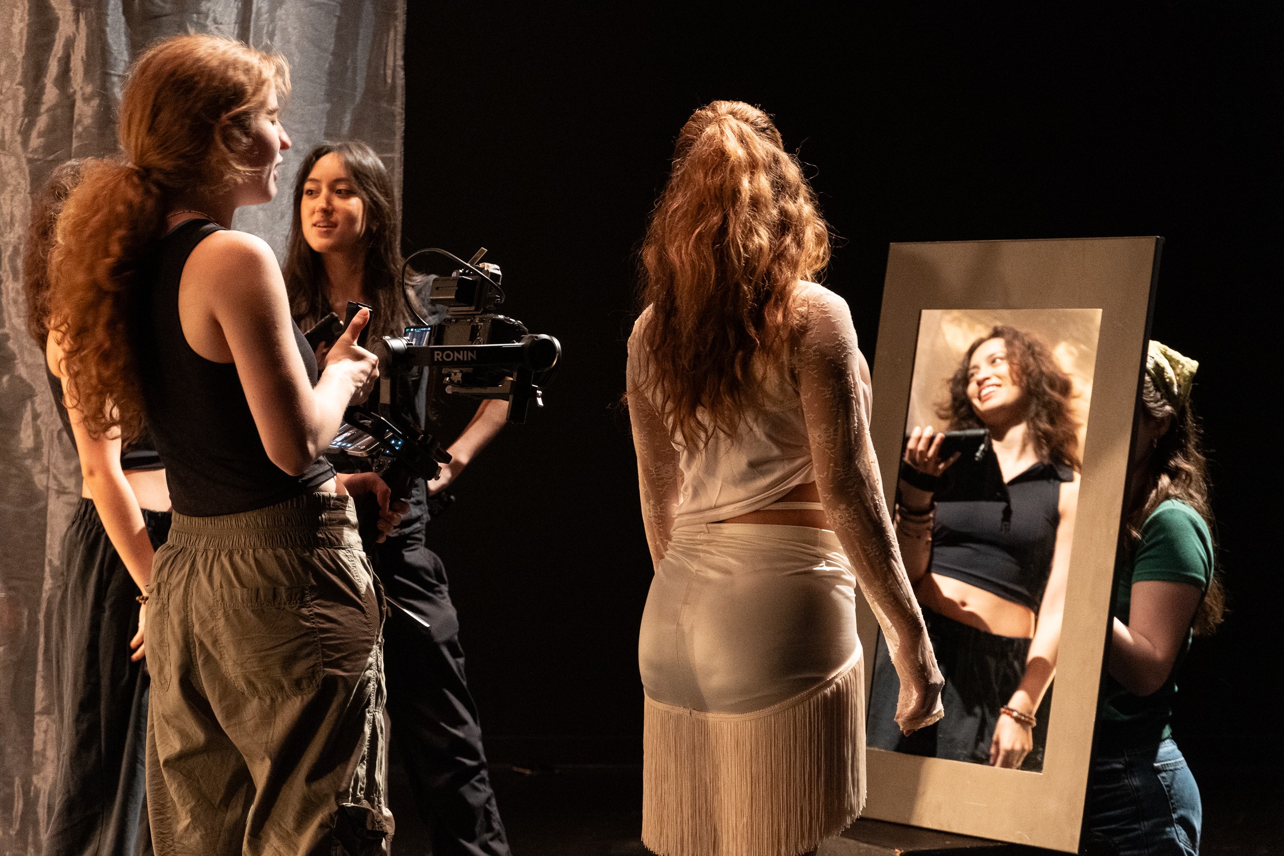 Four women on stage, one looking into a mirror, with a dark background and stage lighting.