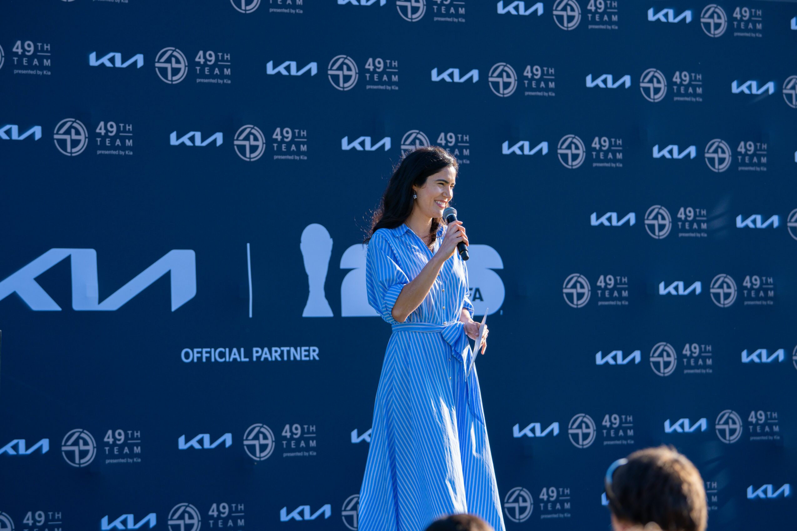Woman in blue dress speaking into microphone on stage with blue background and logos.