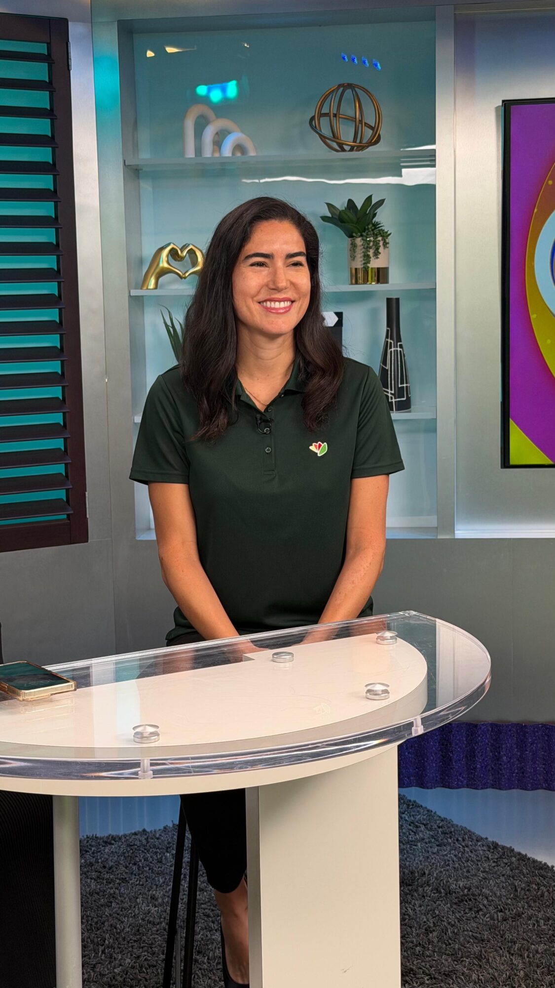 Woman with dark hair smiling, standing behind a transparent desk in a studio with shelves and colorful decor