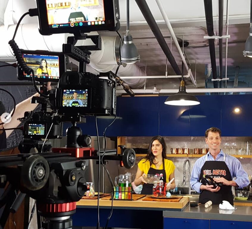 Two people sit at a table with cameras and lighting equipment filming in a modern kitchen studio.