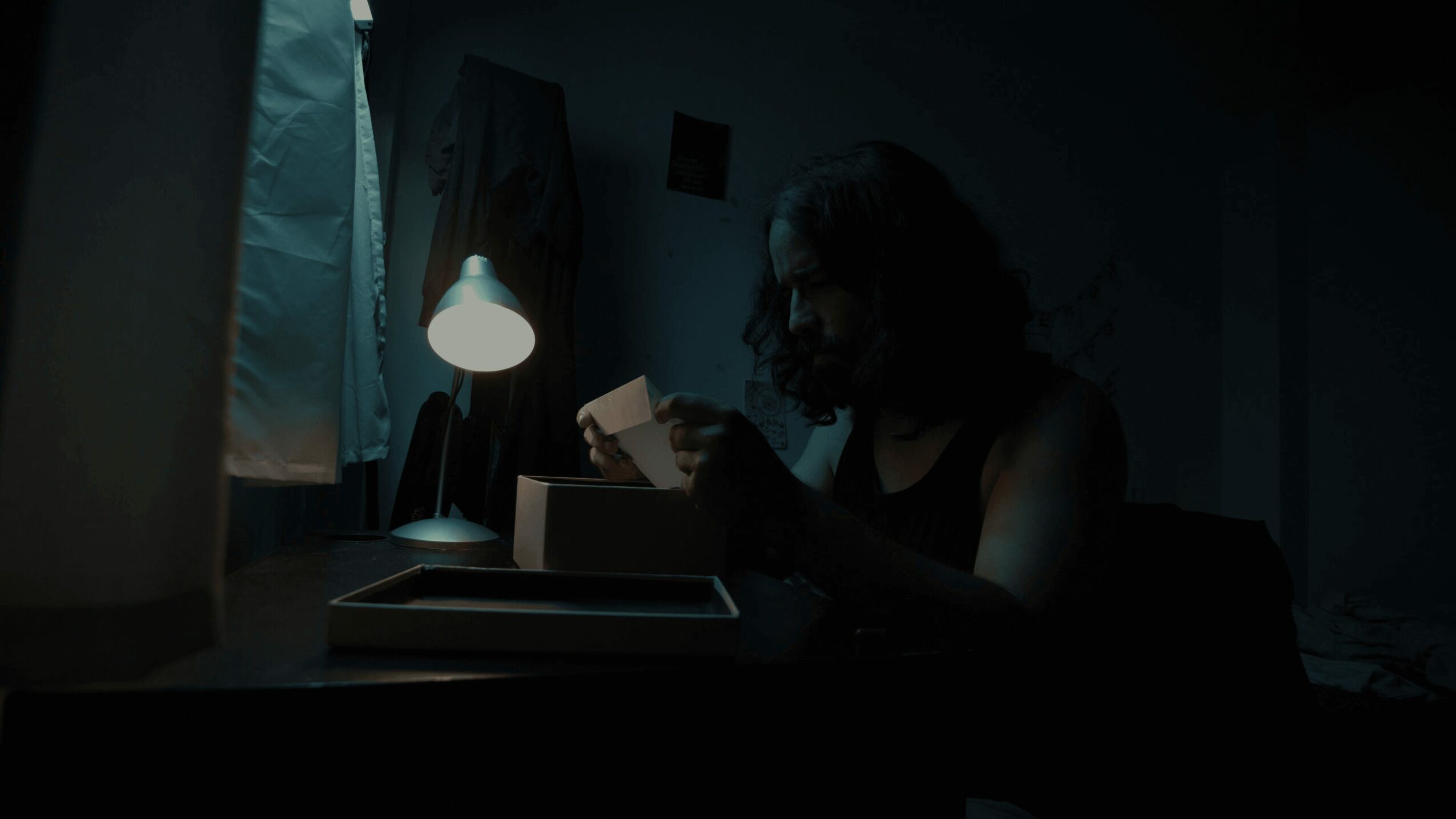 Person sitting at a desk in a dark room, illuminated by a small desk lamp, holding a cup.
