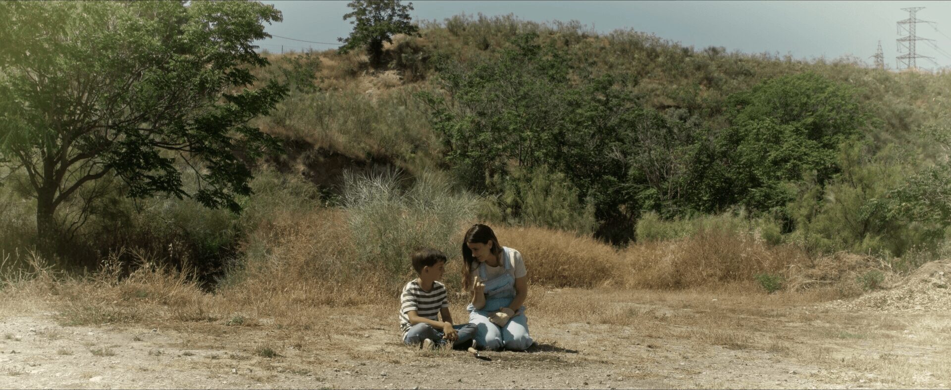 Two children sitting on the ground outdoors, with trees and hills in the background.