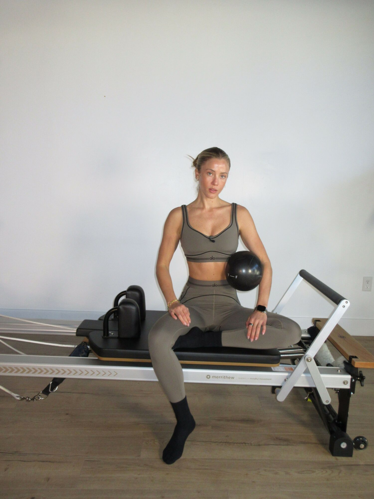 Woman in workout clothes sitting on exercise equipment holding a black ball, in a room with white wall and wooden floor.