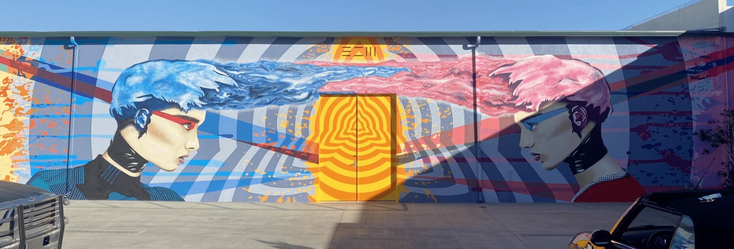 Colorful mural with two stylized faces, abstract patterns, and a central yellow-orange element, painted on a wall.