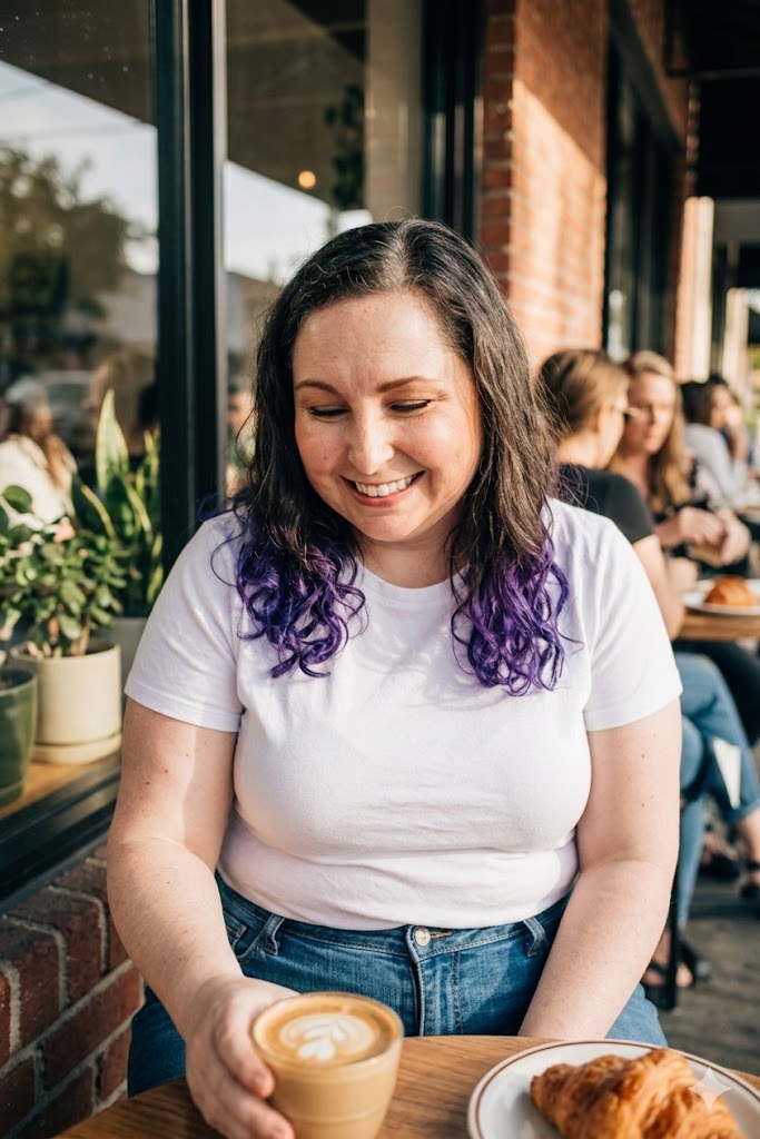Smiling woman with dark and purple hair sitting at a table outside a cafe, with food and drinks, sunlight, and other people in background.