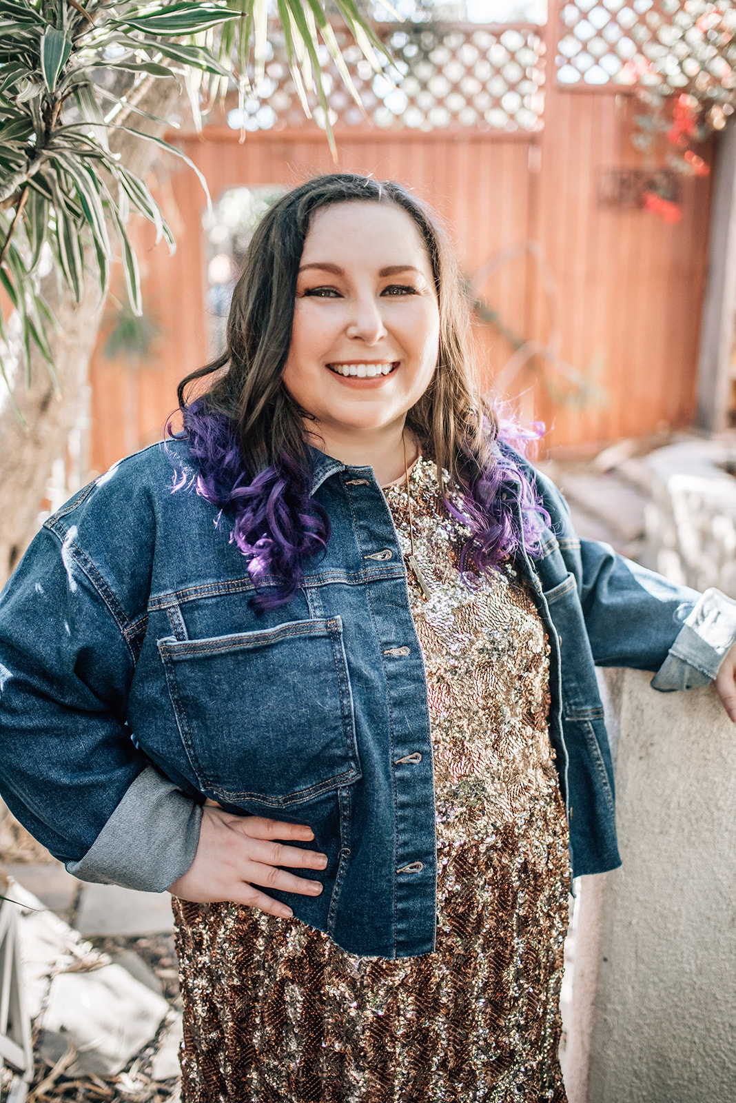 Smiling woman with dark hair and purple tips, wearing a denim jacket and patterned dress, outdoors with plants and a wooden fence.