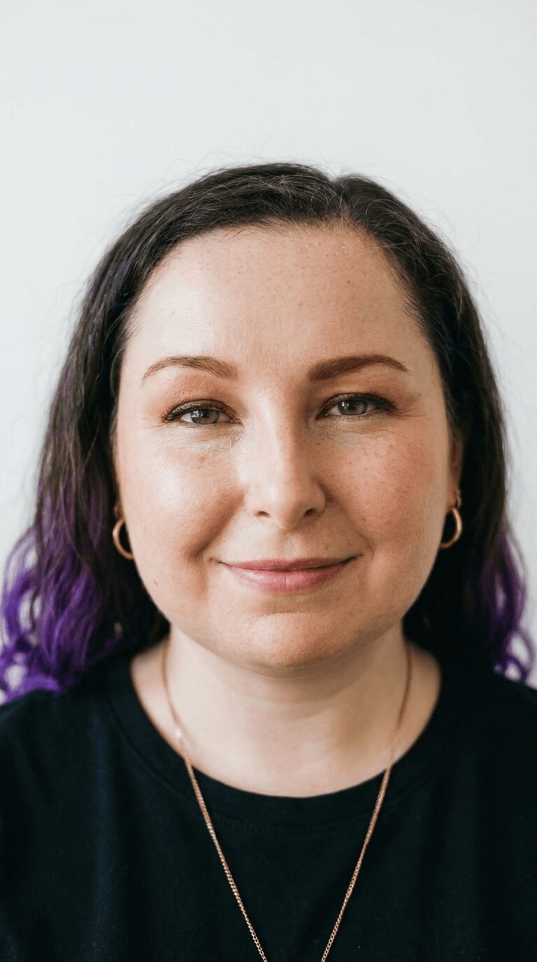 Portrait of a woman with dark hair and purple tips, smiling, wearing a black top and gold jewelry.