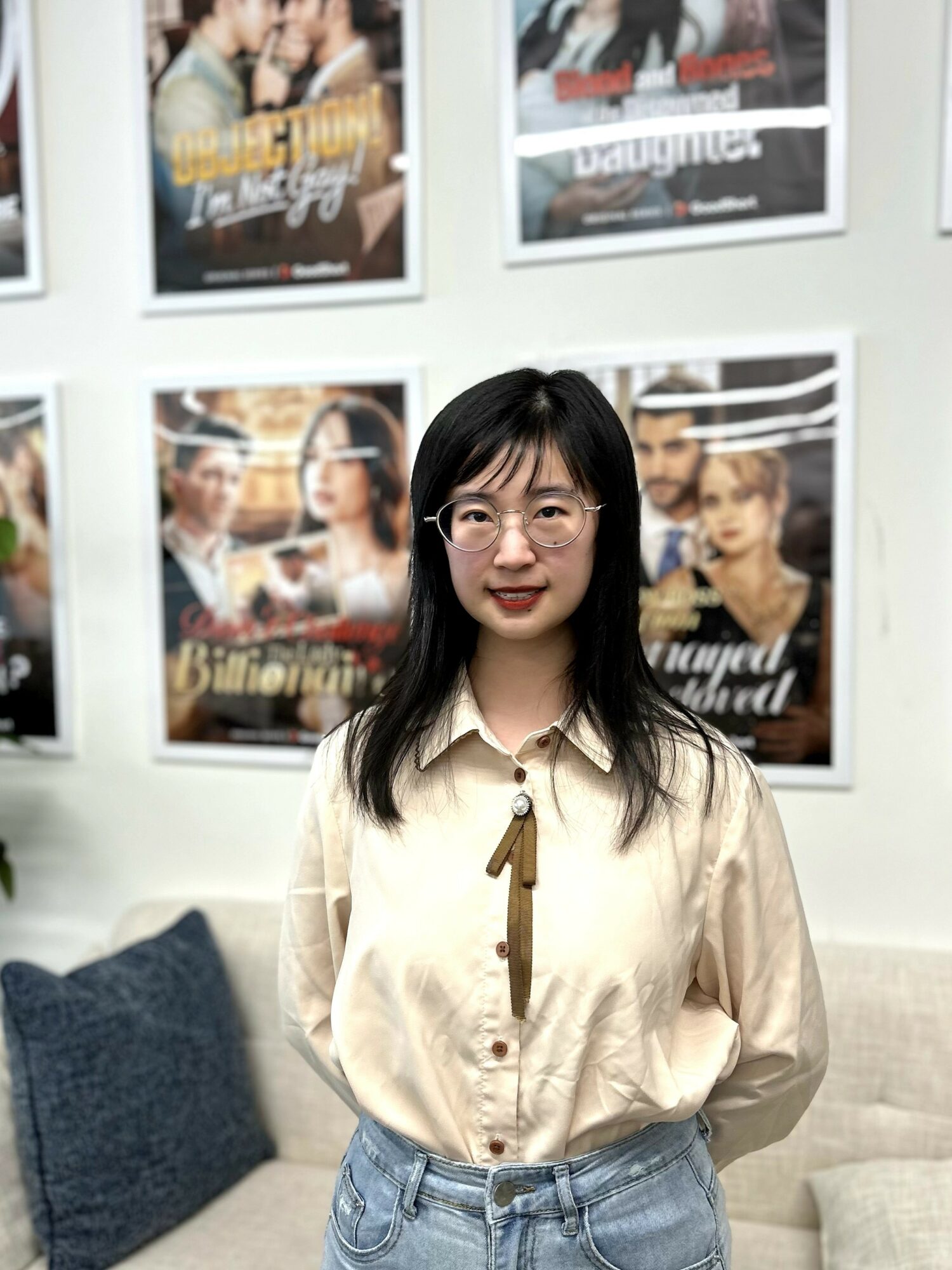 Young woman with glasses and long black hair standing in front of movie posters, wearing a beige shirt and jeans.