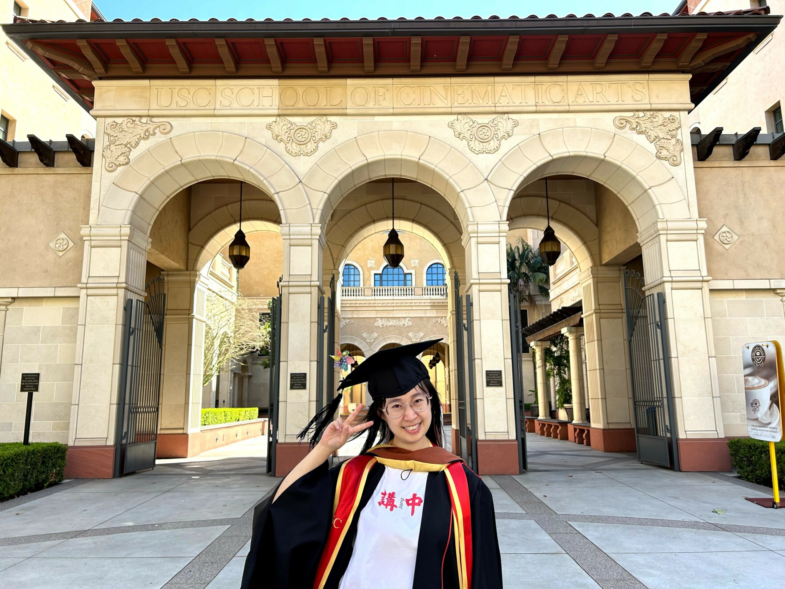 Young woman in graduation gown and cap smiling and making a peace sign in front of an ornate archway.