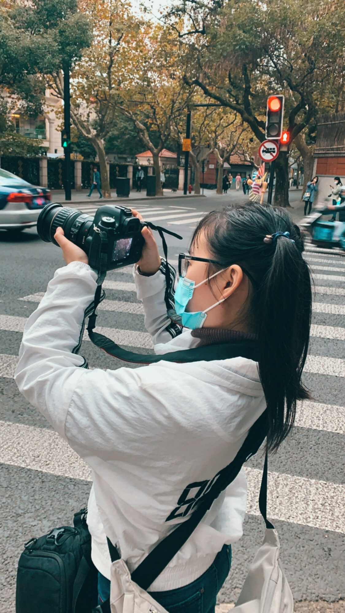 Woman with glasses and face mask holding a camera at a crosswalk in an urban setting.
