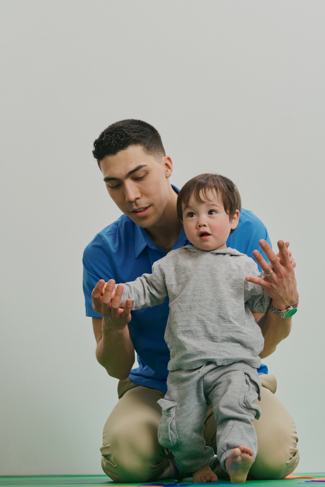 Adult holding a young child with outstretched arms, both sitting on the floor against a plain background.