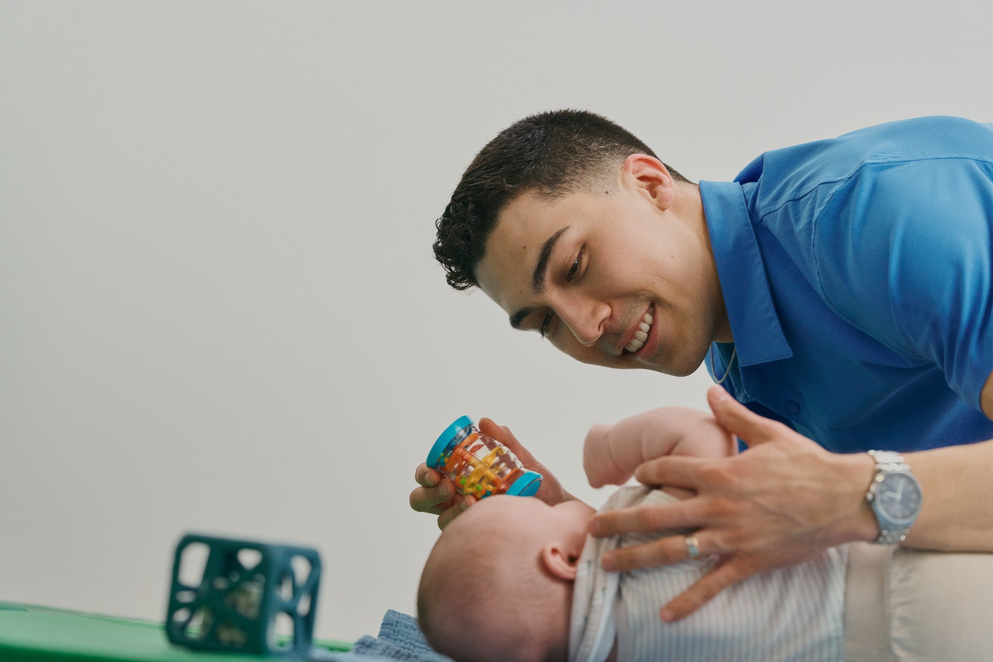 Young man smiling and talking to a baby lying on a surface, with a smartphone nearby, against a plain background.