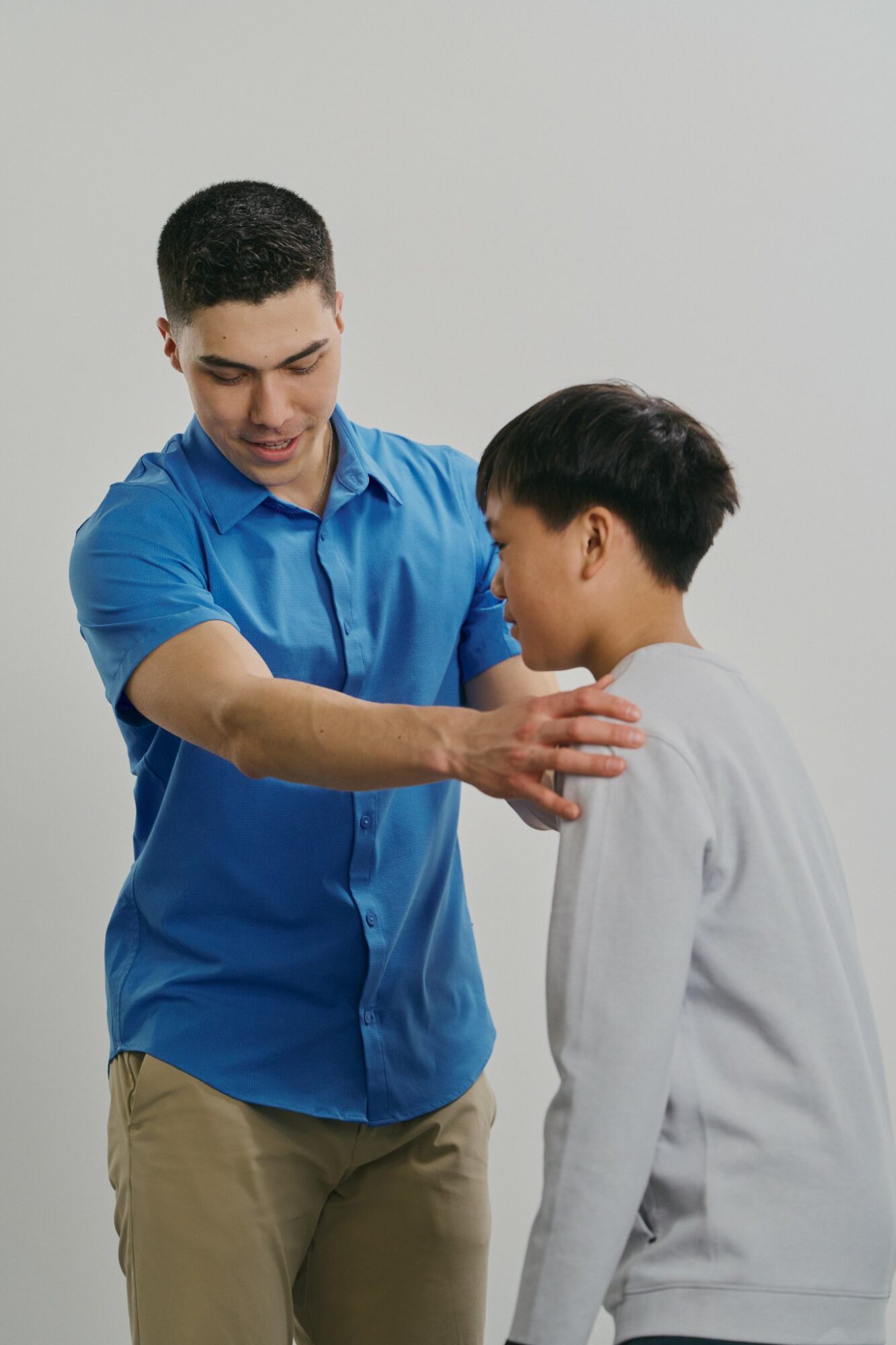 Adult man in blue shirt gently touches a young boy's shoulder, both with neutral expressions, against a plain background.