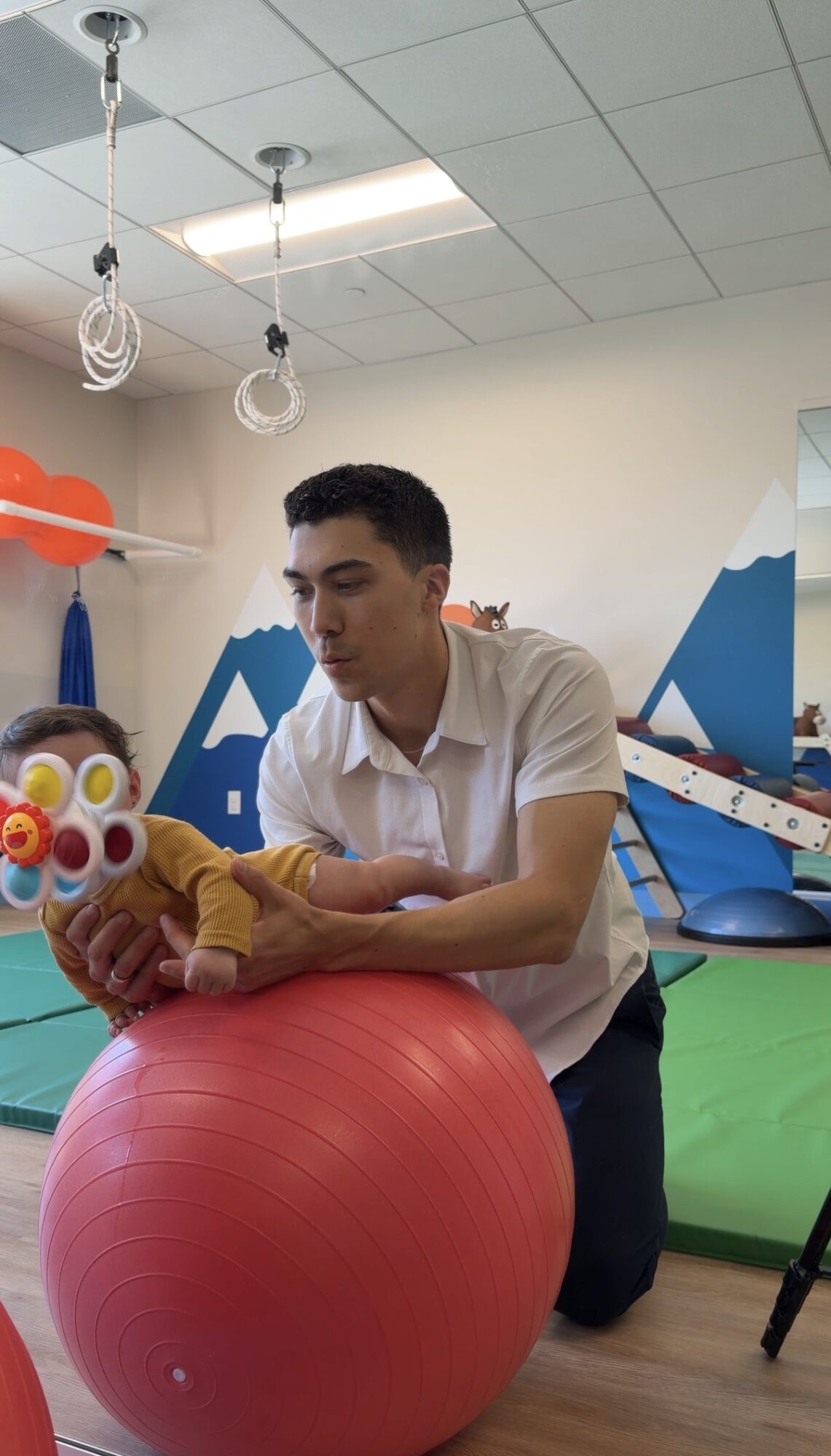 Young man kneeling on the floor holding a plush toy, with gym equipment and a blue wall in the background.