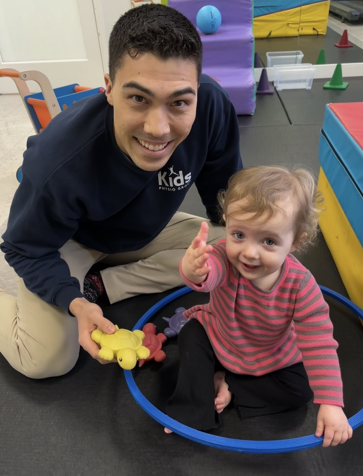 Young man and girl sitting on the floor, smiling, with colorful play equipment around them.