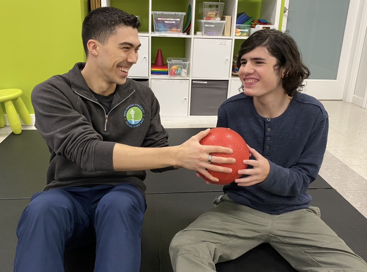 Two people sitting on a black couch, smiling, holding a red ball together in a room with colorful shelves.