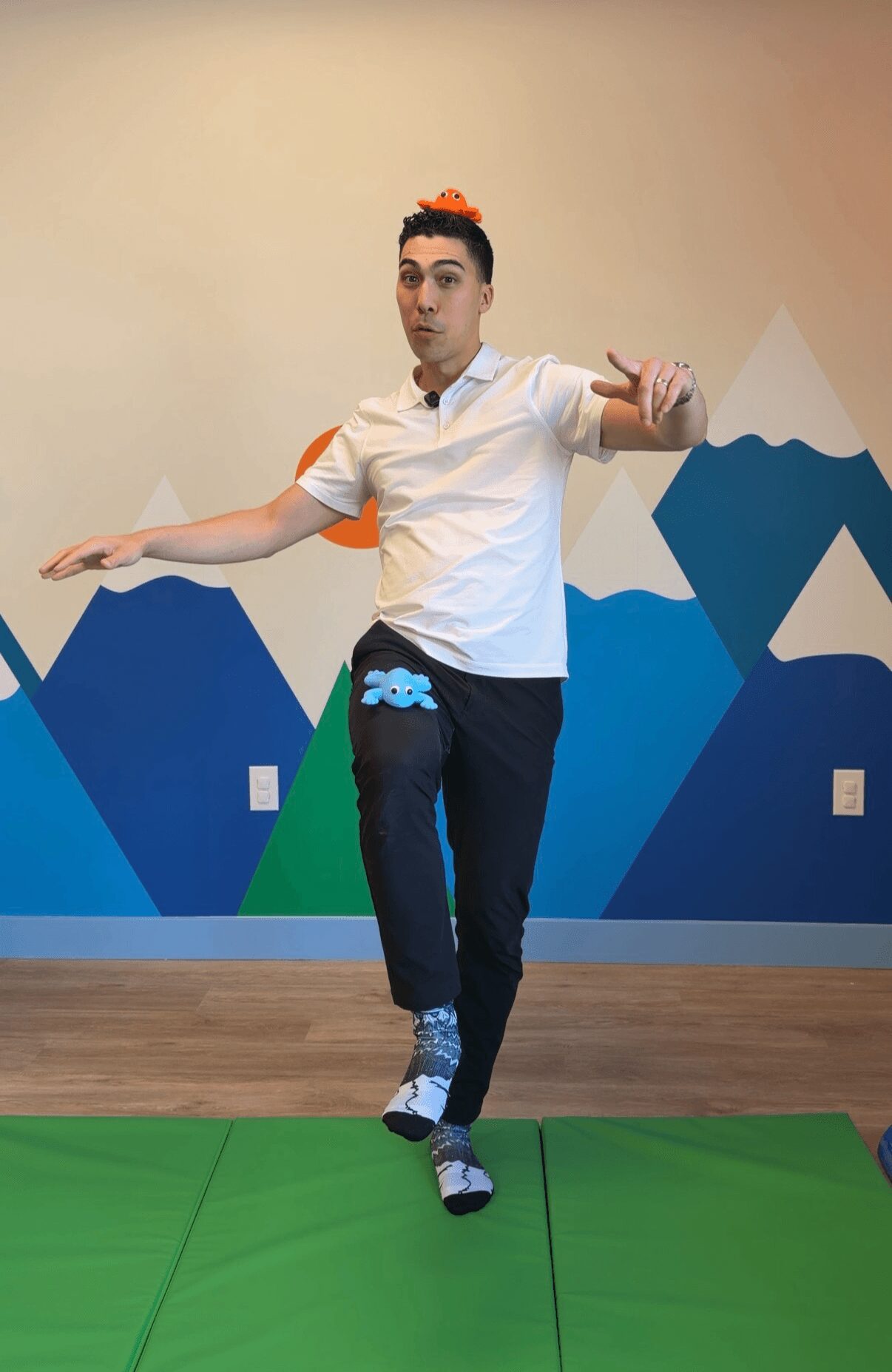 Person standing on green mat with arms extended, in front of a mountain-themed wall, performing a dance move.