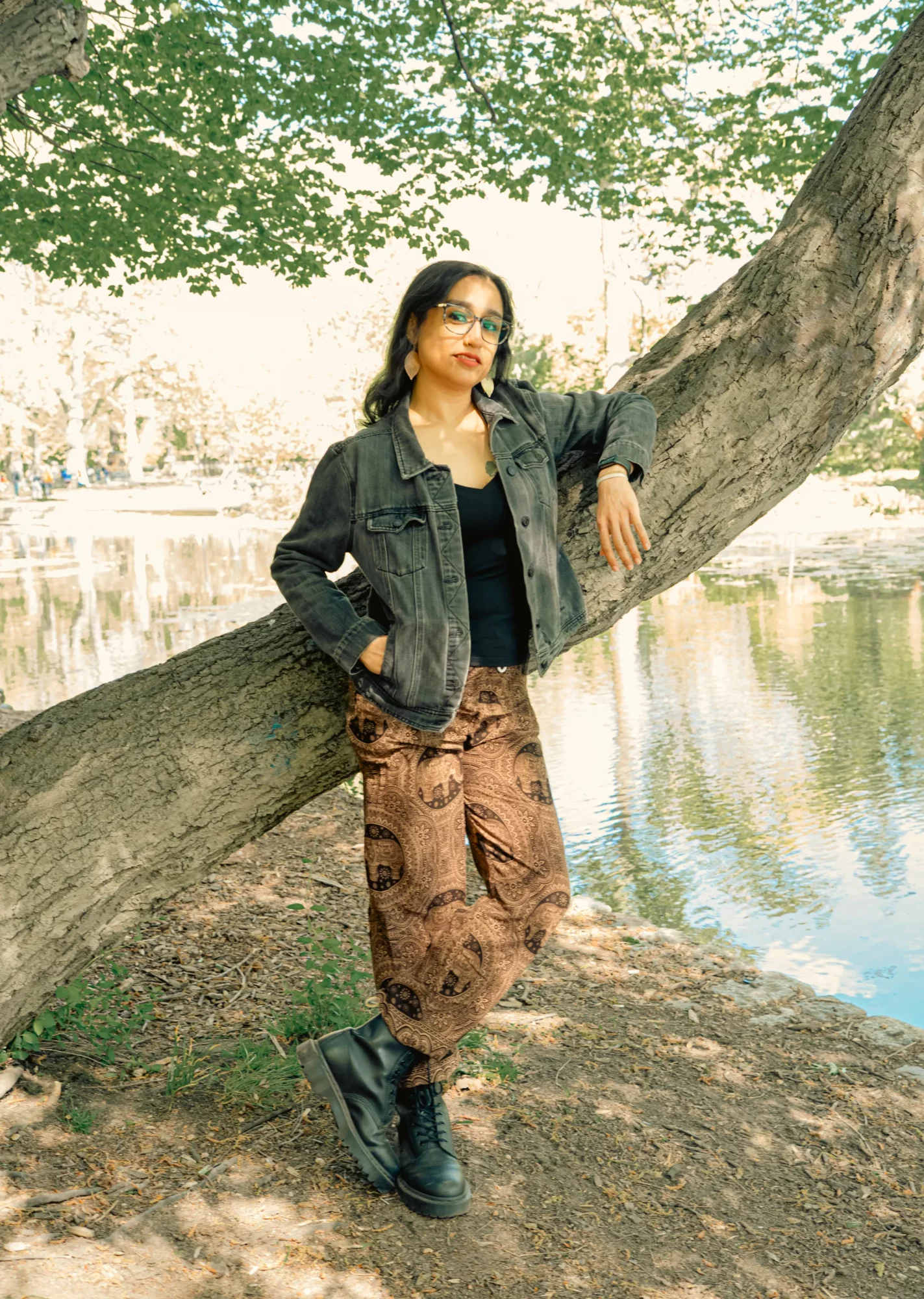 Woman leaning against a tree near a body of water, wearing glasses, a denim jacket, and patterned pants.