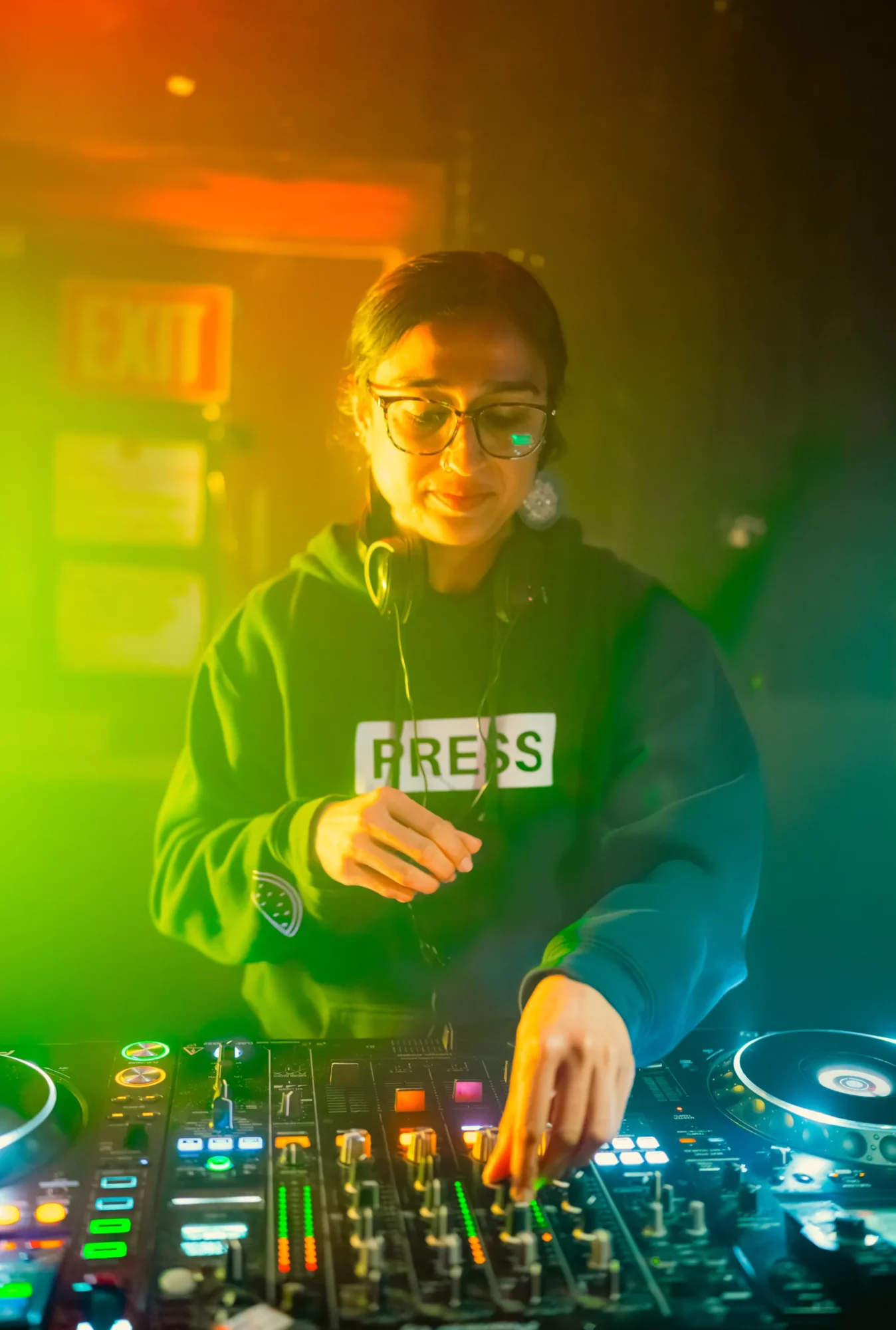 Person wearing glasses and a hoodie with 'PRESS' label, operating DJ equipment in colorful lighting.