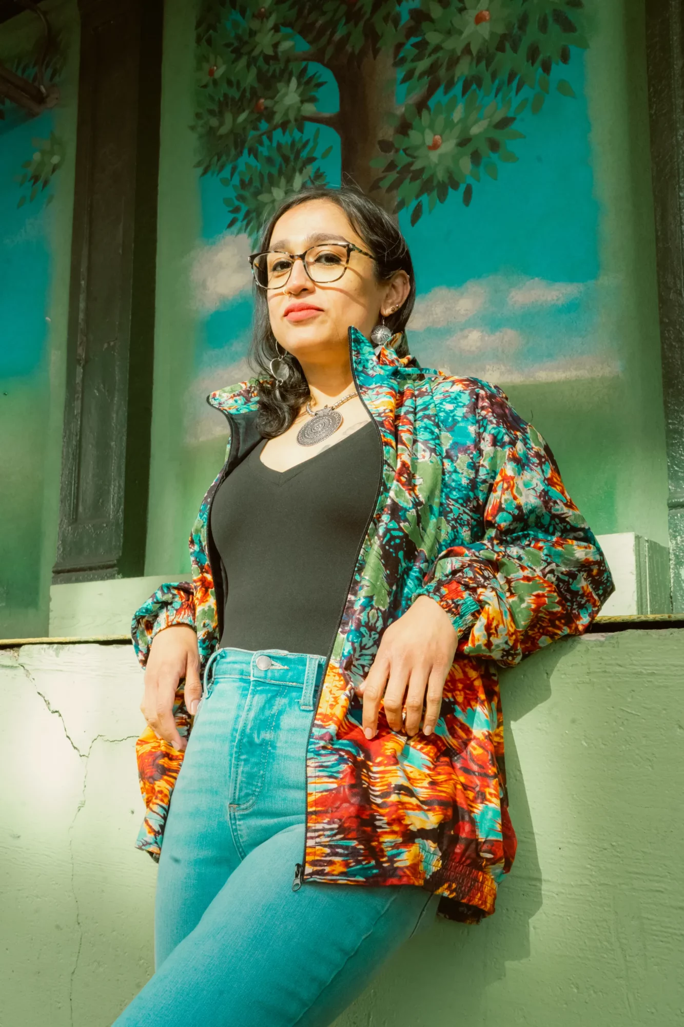 Woman with glasses and dark hair, wearing a colorful jacket and black top, standing outdoors near a painted wall.