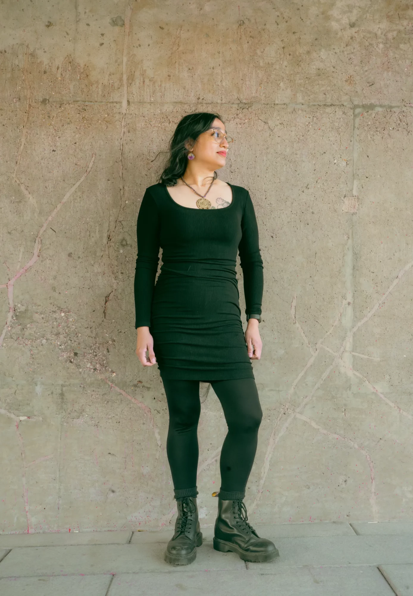 Woman in black dress and boots standing against a textured wall, looking to her left.