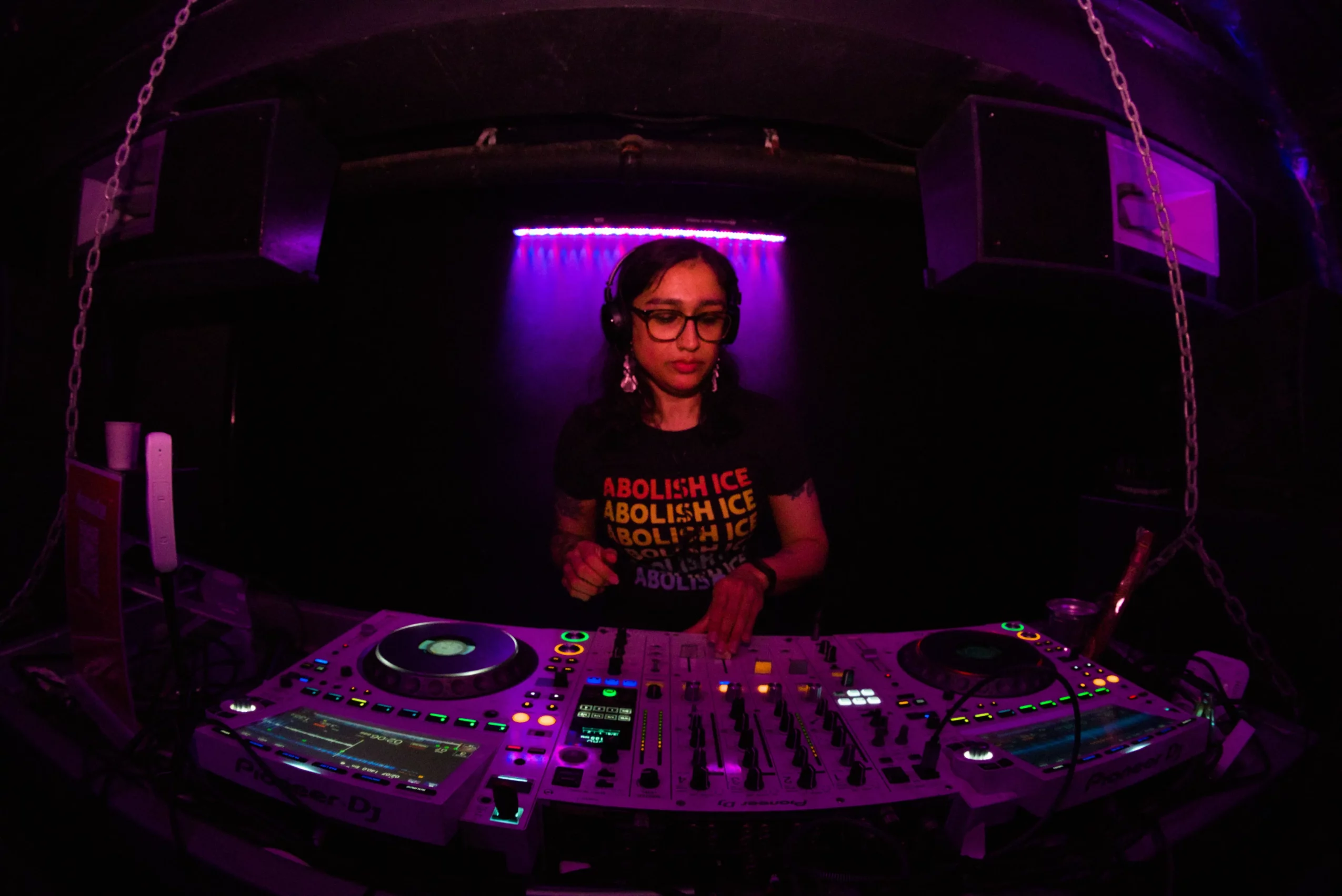 Female DJ wearing glasses and a black shirt with yellow text, standing behind a DJ mixer in a dark room with purple lighting.