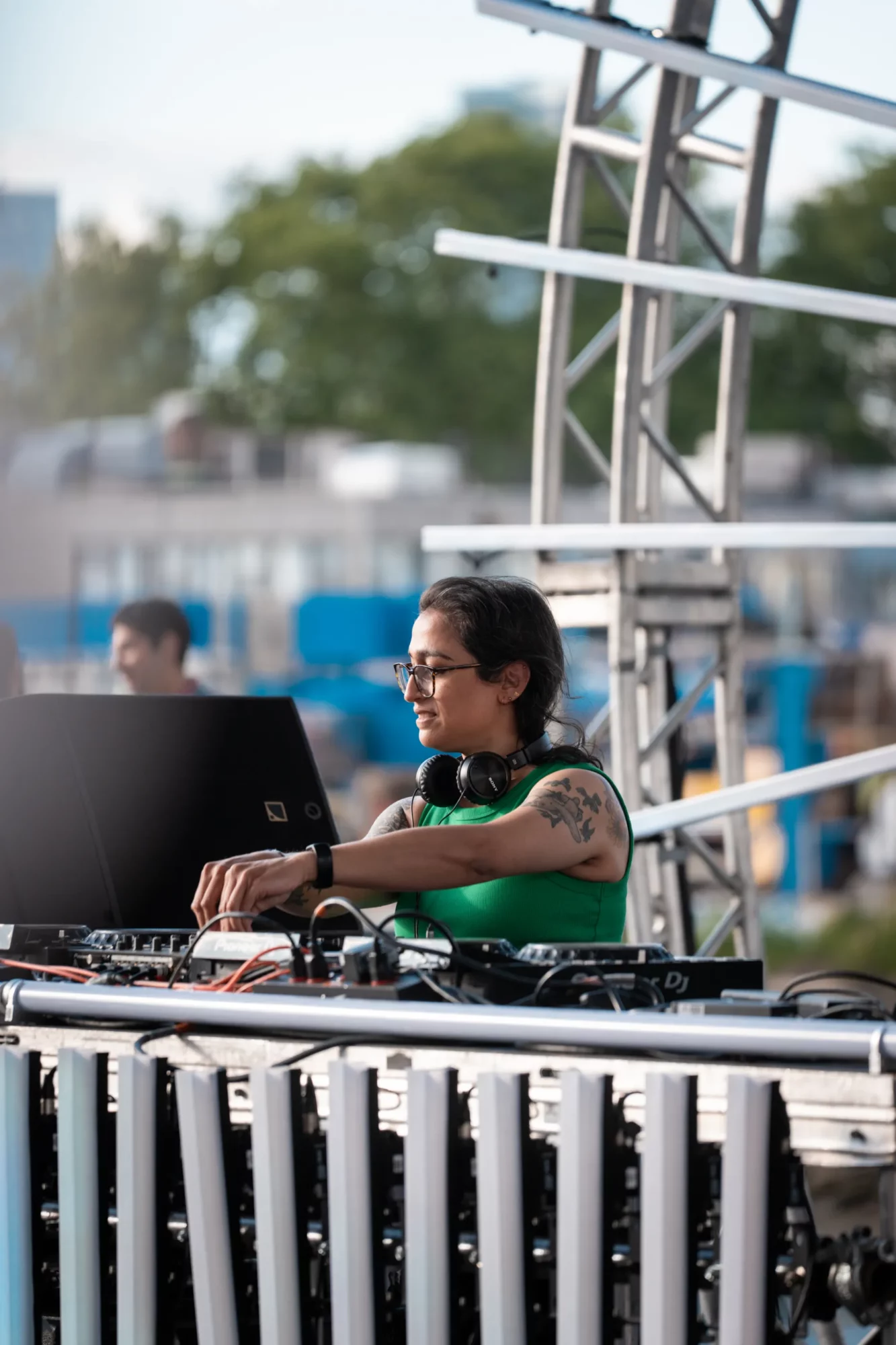Female DJ wearing glasses and headphones, operating DJ equipment outdoors with trees and structures in background.
