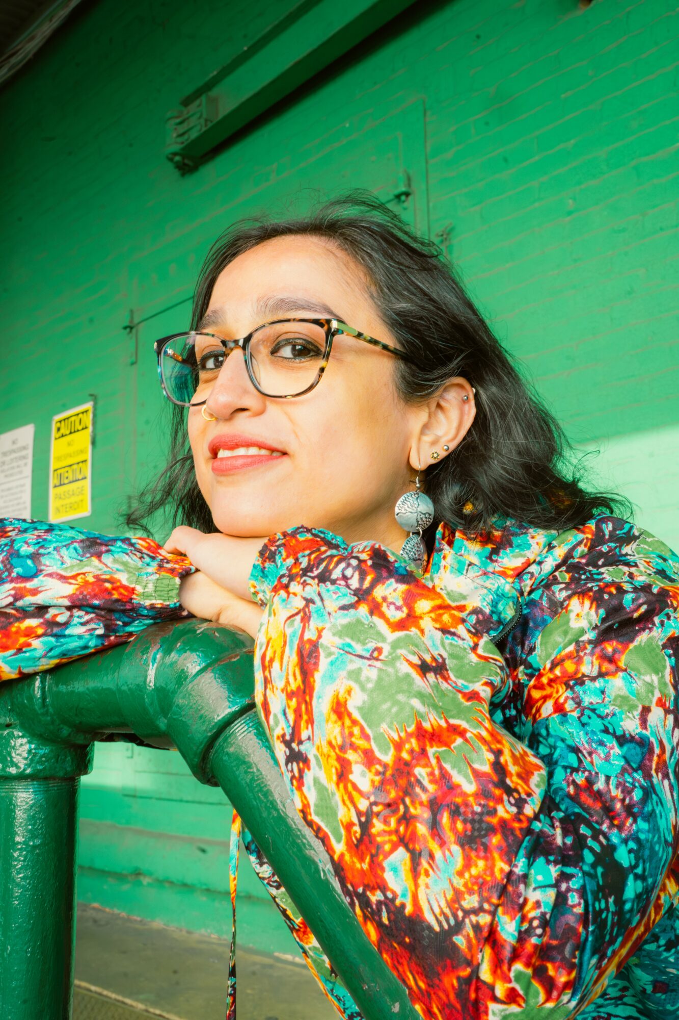 Woman with glasses and earrings leaning on a green railing, smiling, in front of a green wall.