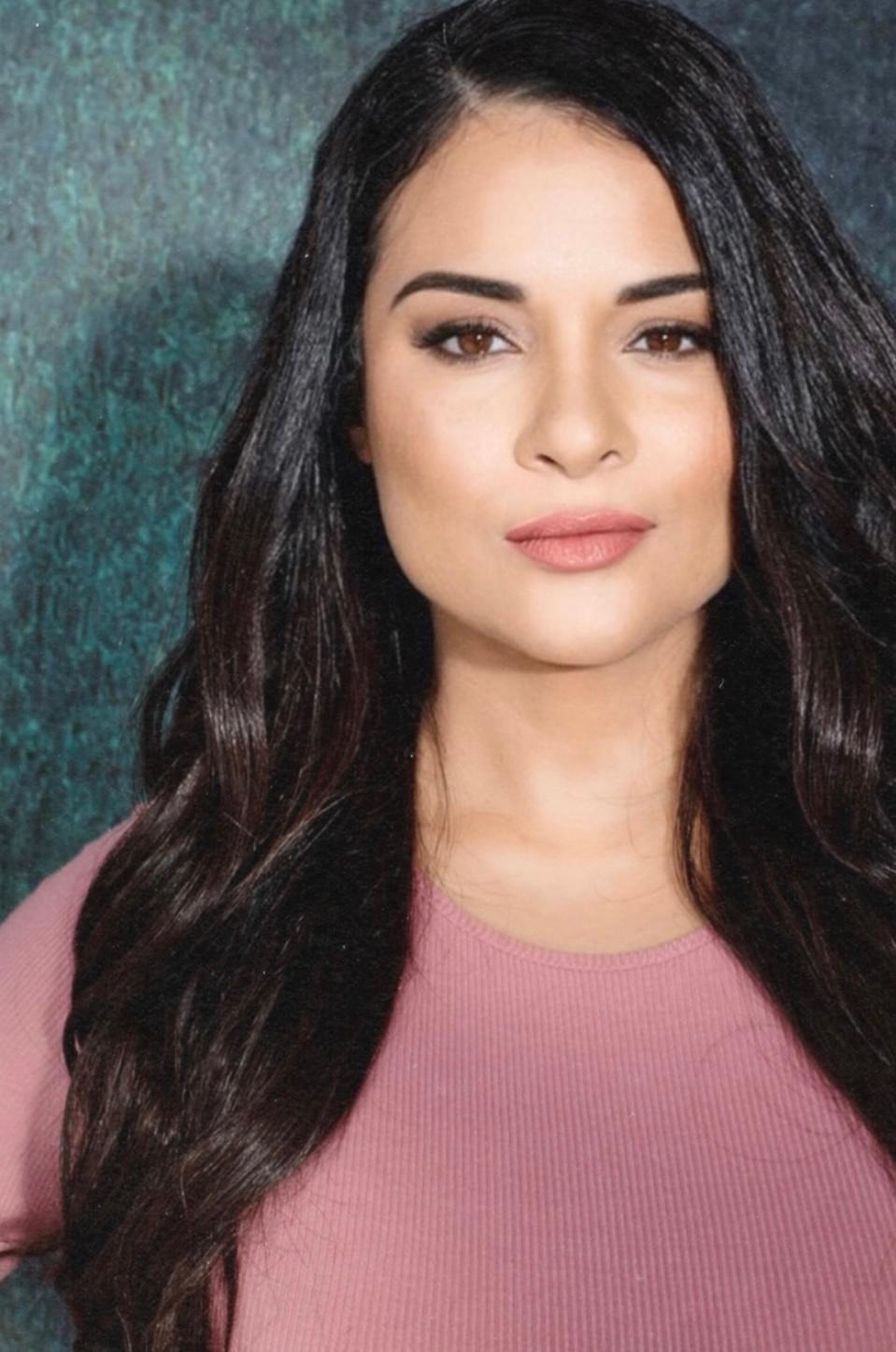 Young woman with long dark hair and light makeup, wearing a pink top, looking at the camera.