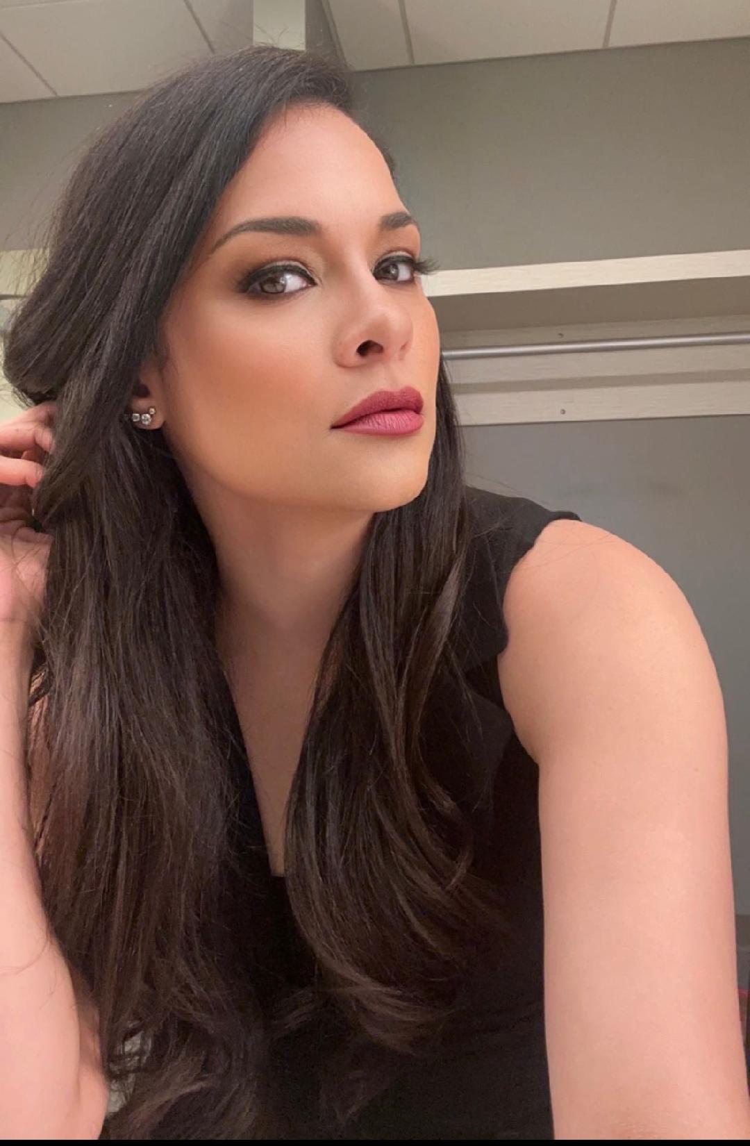 Woman with long dark hair and makeup, wearing a black sleeveless top, looking at the camera.