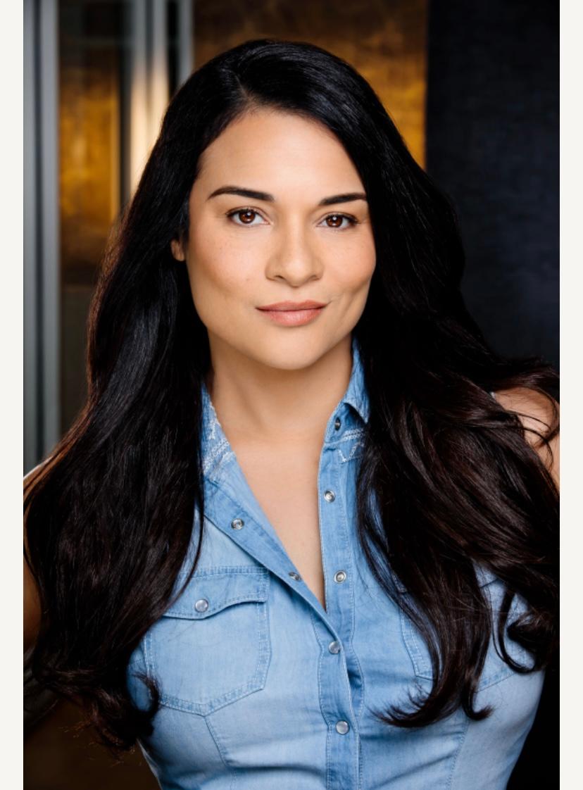 Woman with long dark hair wearing a blue denim shirt, smiling softly, indoors with warm lighting.