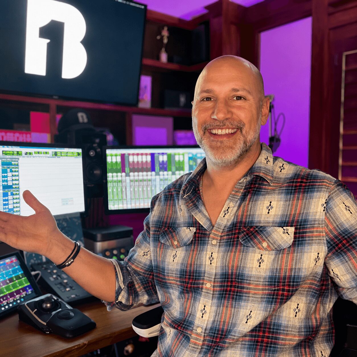 Man smiling in a recording studio with audio equipment and screens, wearing a plaid shirt.
