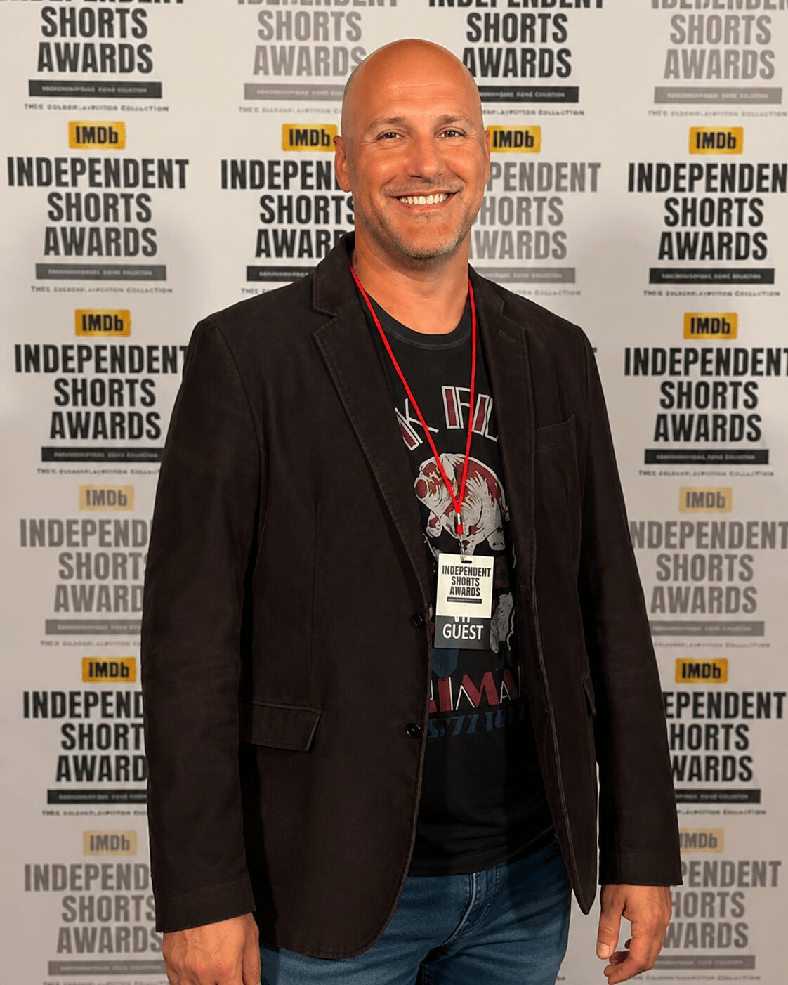 Smiling man in black blazer and T-shirt standing in front of a backdrop with 'Independent Shorts Awards' and 'IMDb' logos.