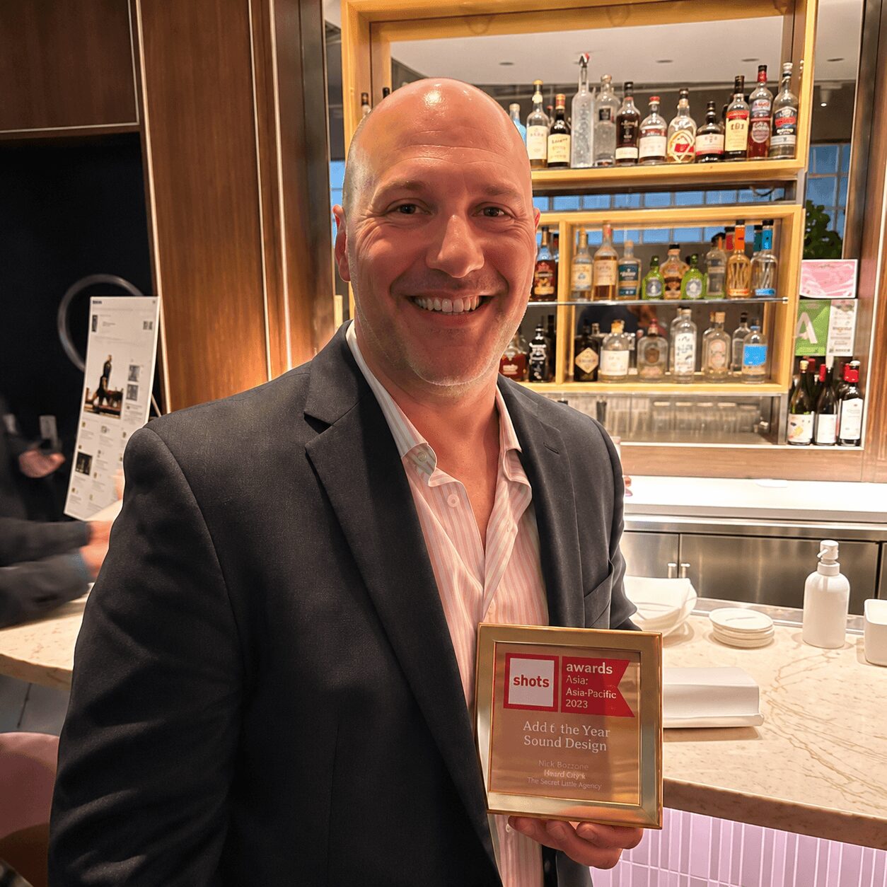 Man in suit smiling, holding a framed award, standing in a bar or restaurant with bottles on shelves behind him.