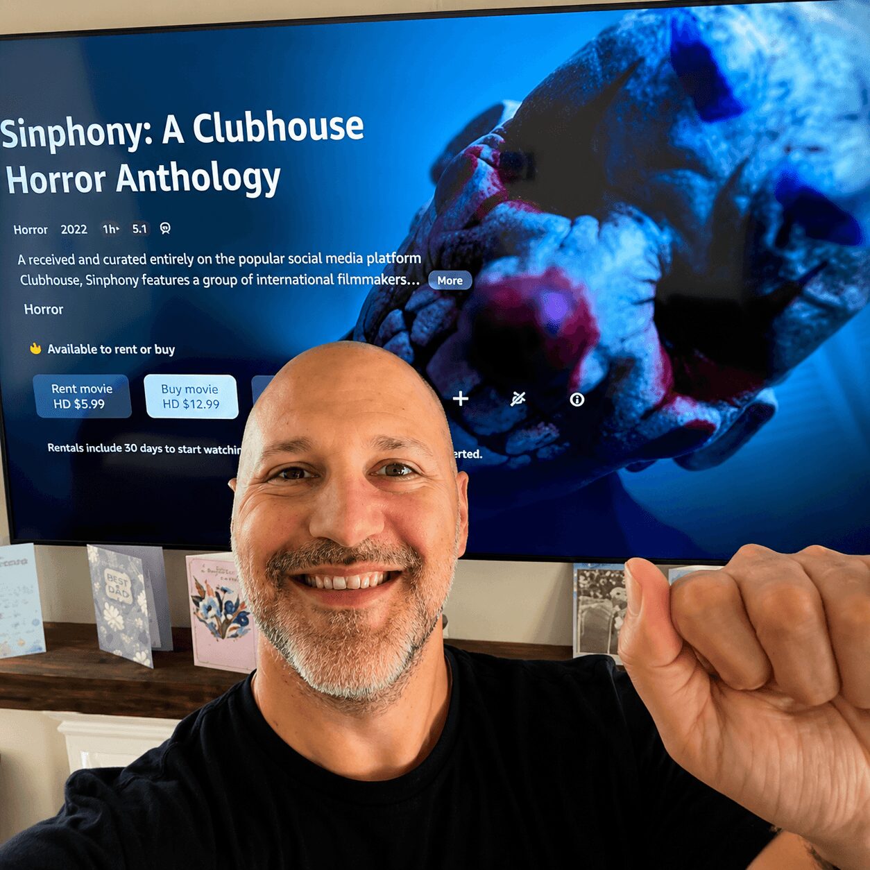 Man smiling in front of a screen displaying a horror movie titled 'Sinphony: A Clubhouse Horror Anthology'.