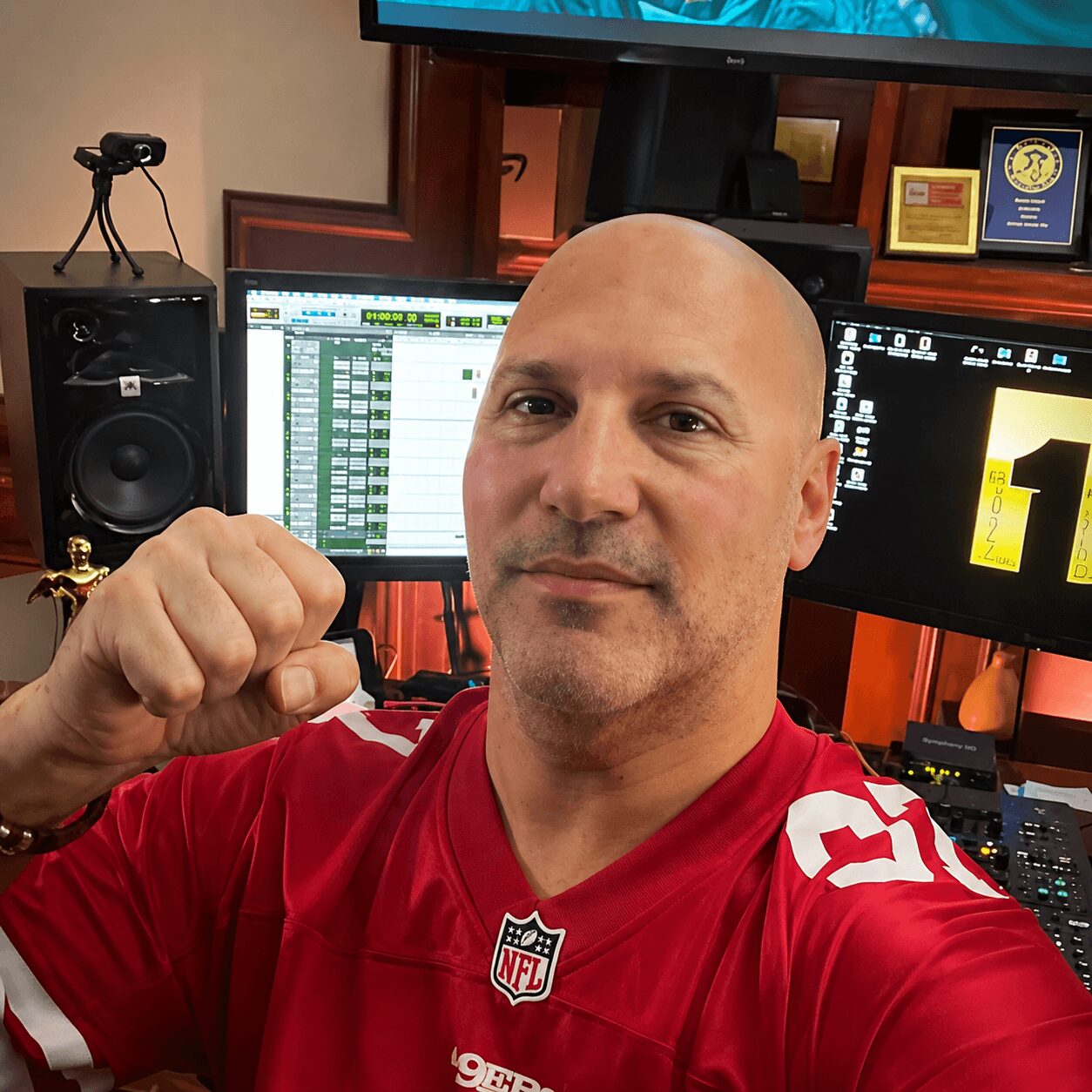 Bald man in a red NFL jersey flexing his arm in a recording studio with multiple monitors and equipment behind him.