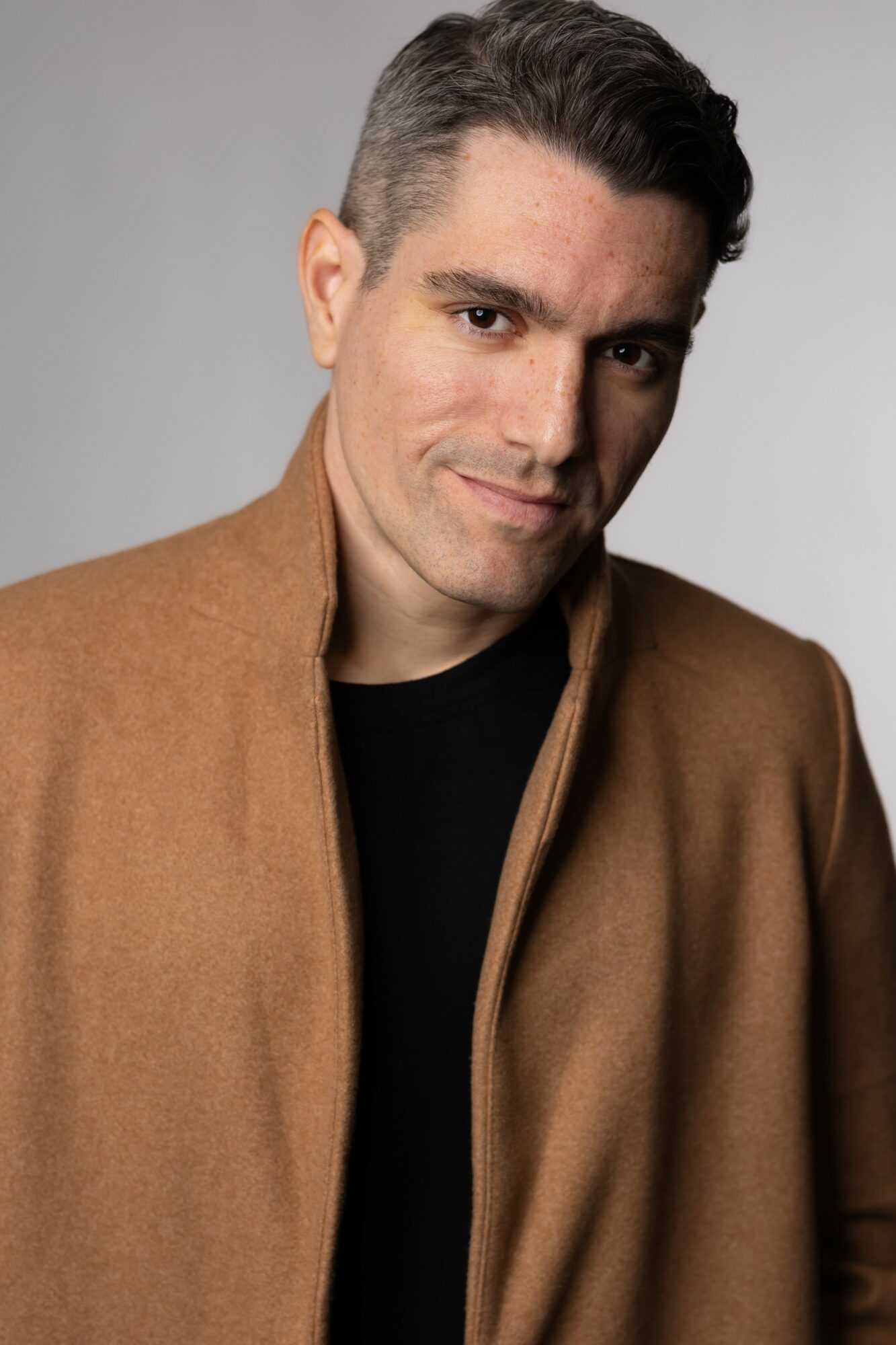 Man with dark hair and a slight smile, wearing a brown coat over a black shirt, against a gray background.